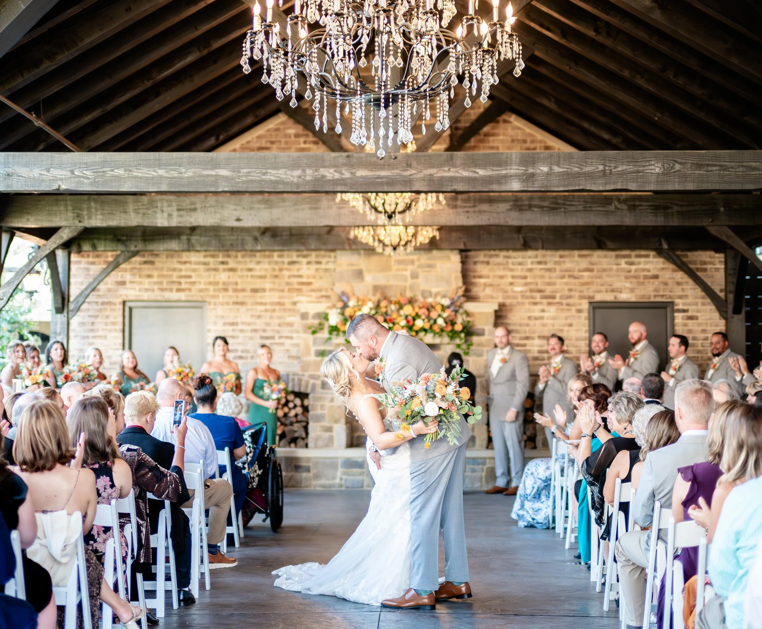 Bride and groom sharing a kiss during their wedding ceremony in a rustic venue with chandeliers, floral arrangements, and guests clapping.