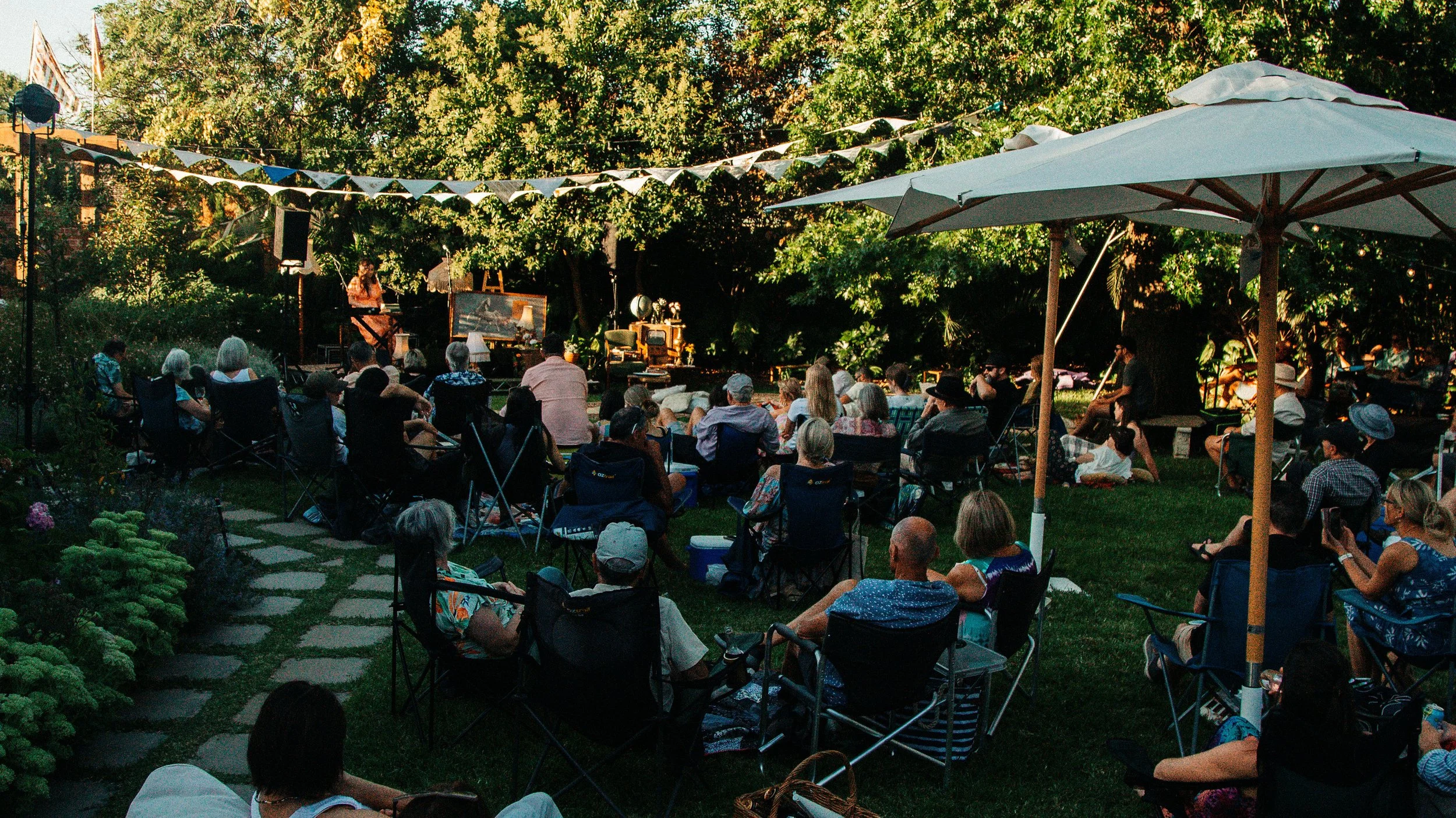 Attendees at the 'Secret Garden Gig' sitting in lawn chairs and sheltered by umbrellas, creating a vibrant crowd.