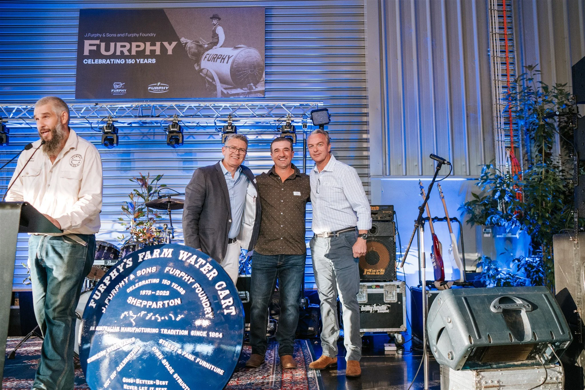 Three individuals posing with a plaque on stage to commemorate Furphy 150 Year Celebration.