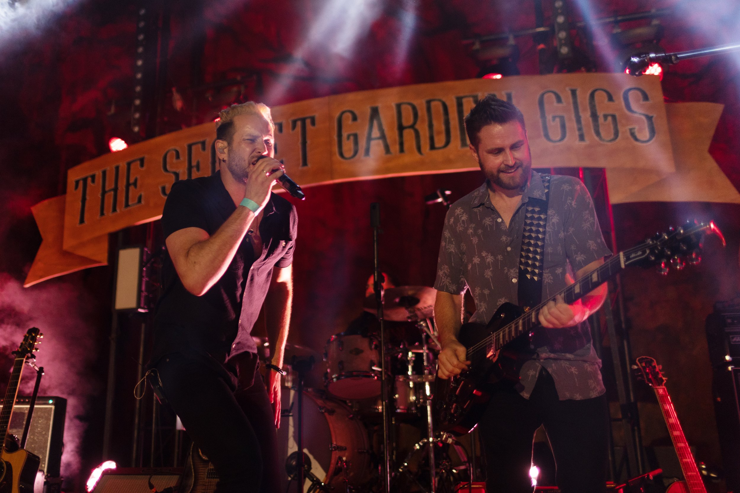 Two men performing on stage in front of a sign that says "The Secret Garden Gigs".