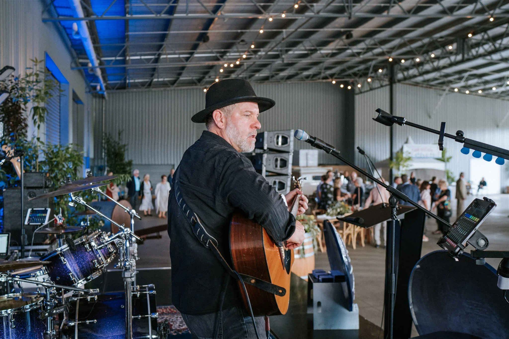 A man in a hat playing an acoustic guitar in a large room at Furphy 150 Year Celebration.