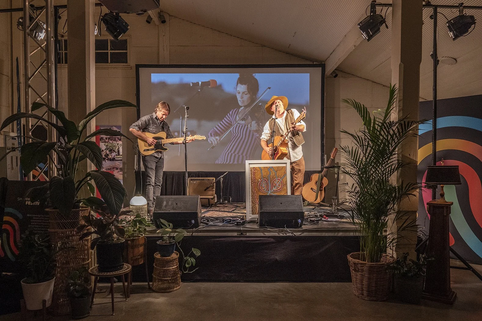 Two musicians performing with guitars, in front of a projection screen, with plants.