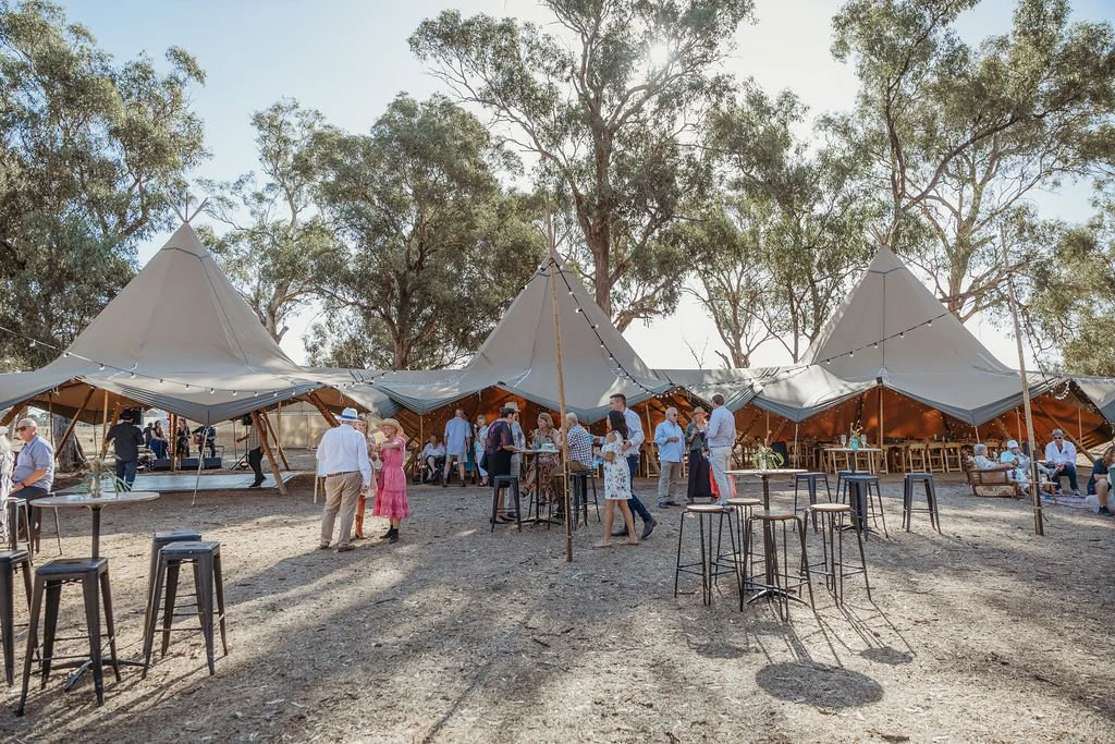 Wedding guests mingling outdoors with spacious tents and greenery in a countryside setting.