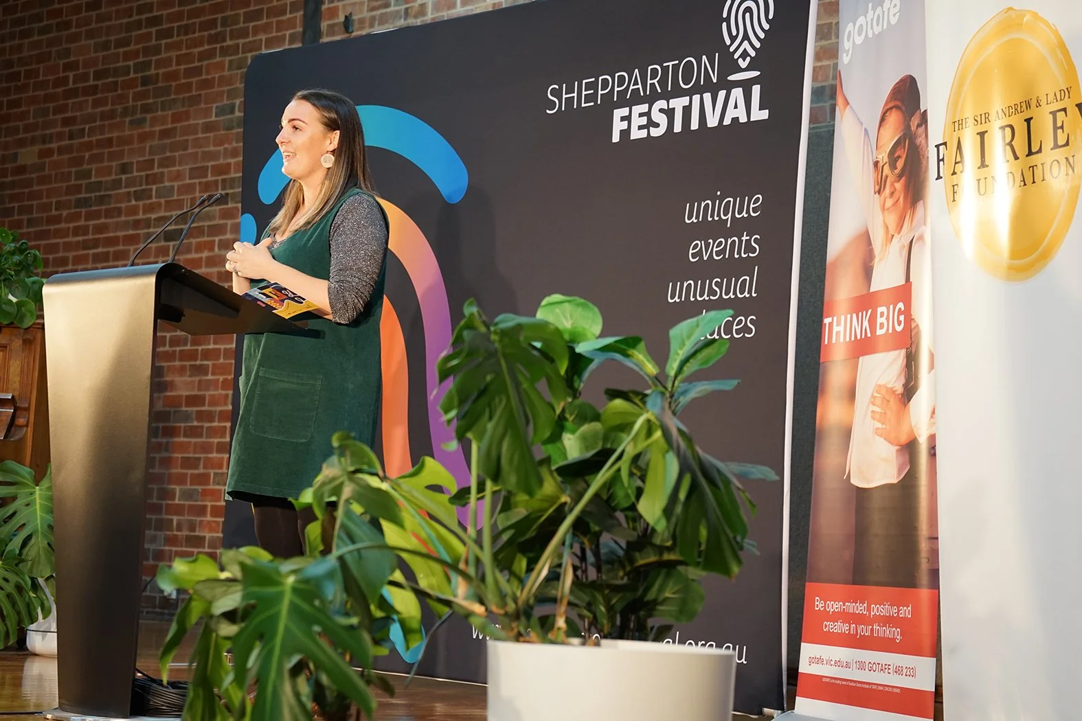 A woman in green giving a speech in front of a sign for the Shepparton Festival.