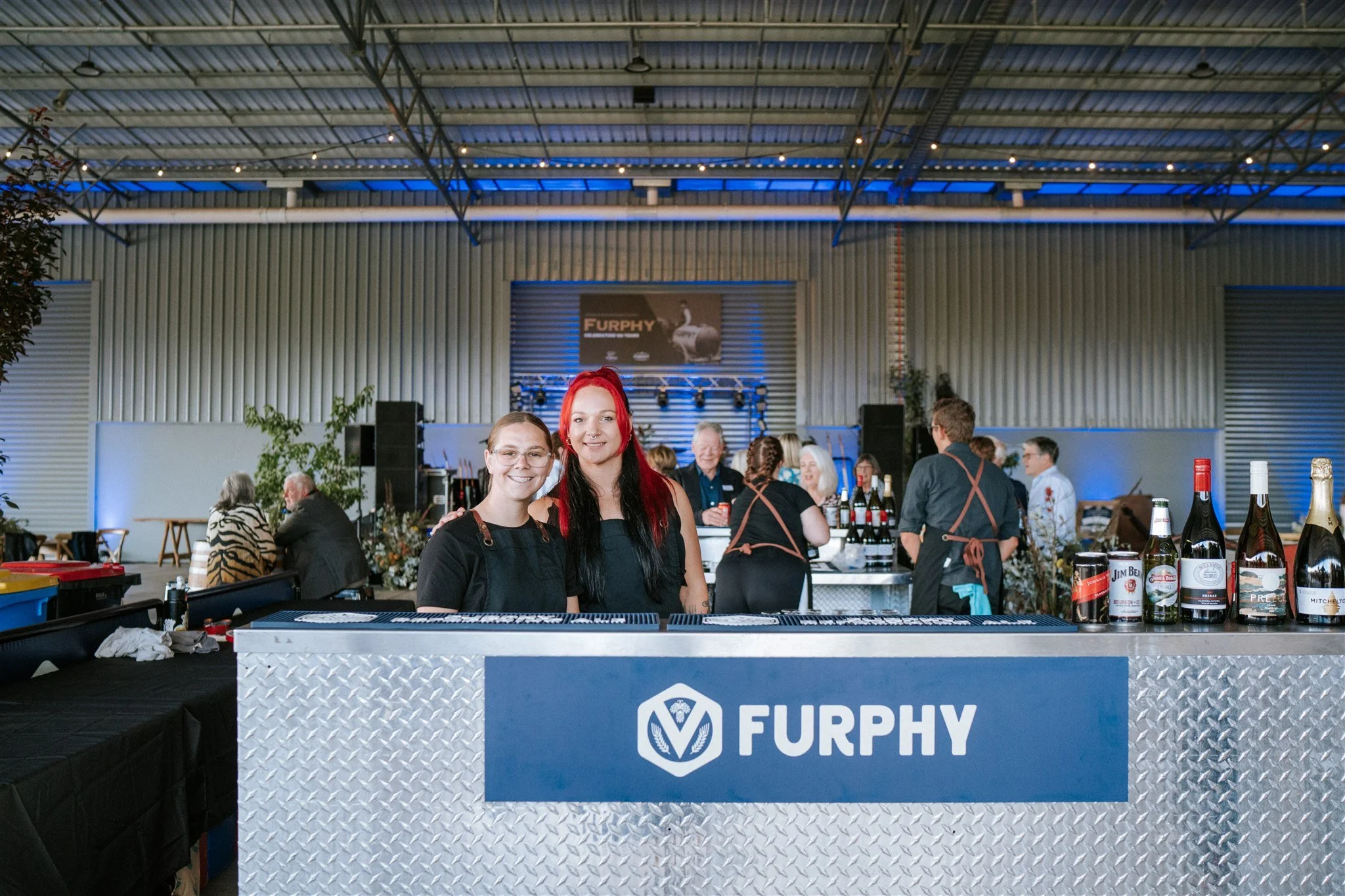 Women standing behind a bar at a wine tasting event for Furphy 150 Year Celebration.