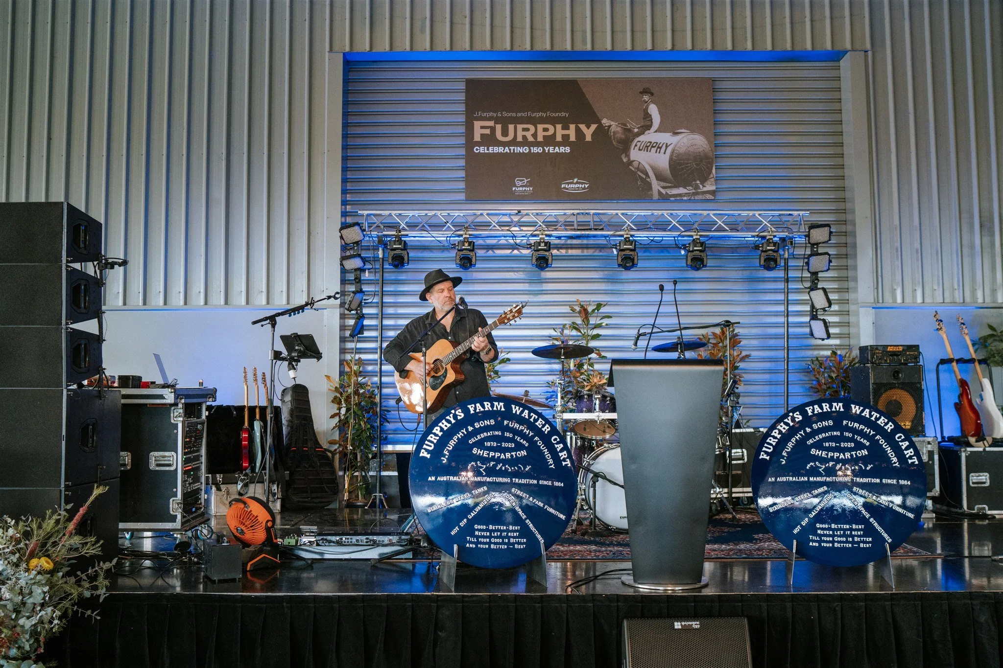 A man playing guitar on stage in front of a blue sign at the Furphy 150 Year Celebration.