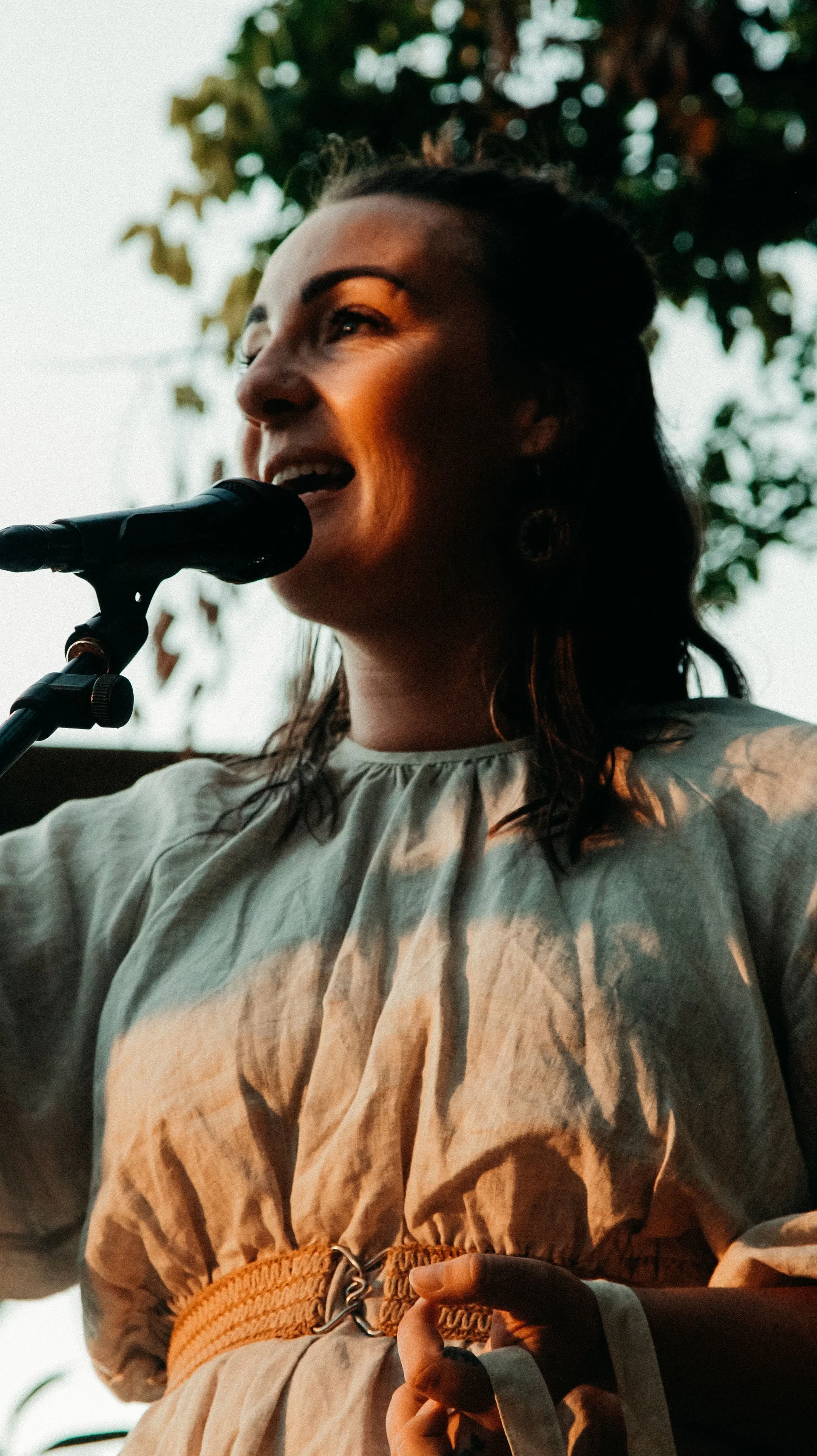 Woman speaks into microphone in front of trees.