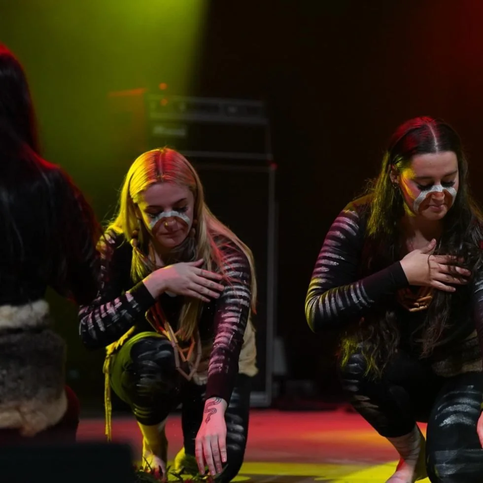 Three women in tribal clothing kneeling on the ground, celebrating Treaty Day Out with traditional attire.