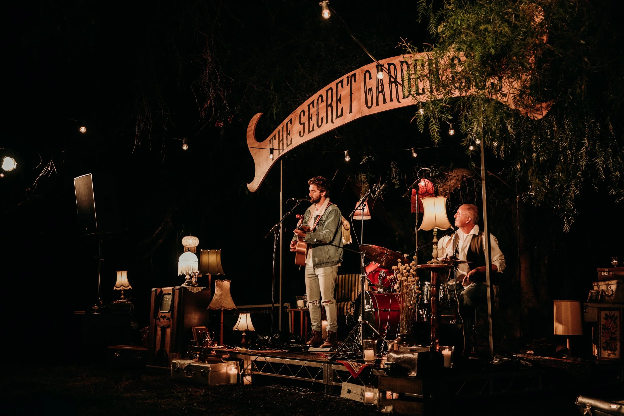 A young man plays guitar, while an older man plays the drums, under a sign for The Secret Garden Gigs.