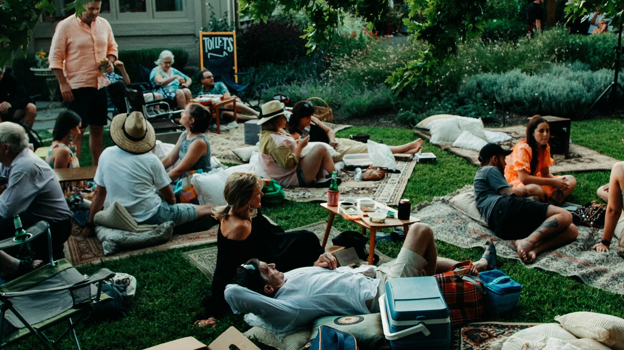 People seated on the green grass in a garden, under the shade of a large tree.