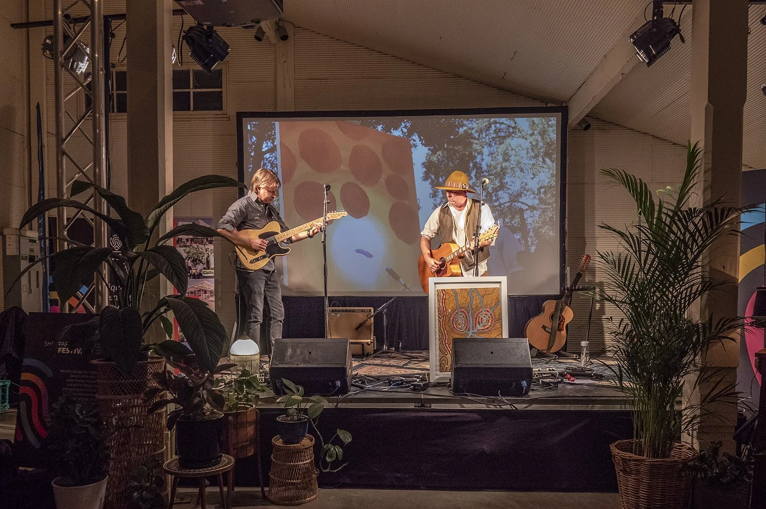 Two musicians performing with guitars, in front of a projection screen, with plants.