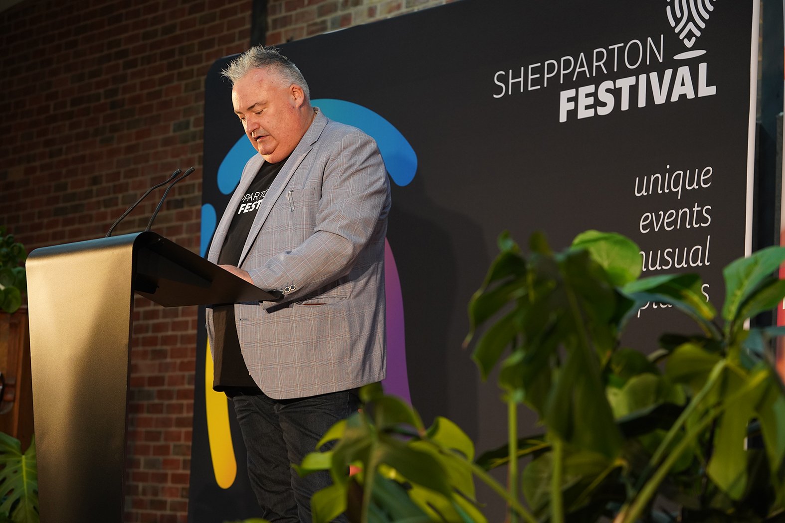 A gentleman in a grey jacket gives a speech in front of a Shepparton Festival sign.
