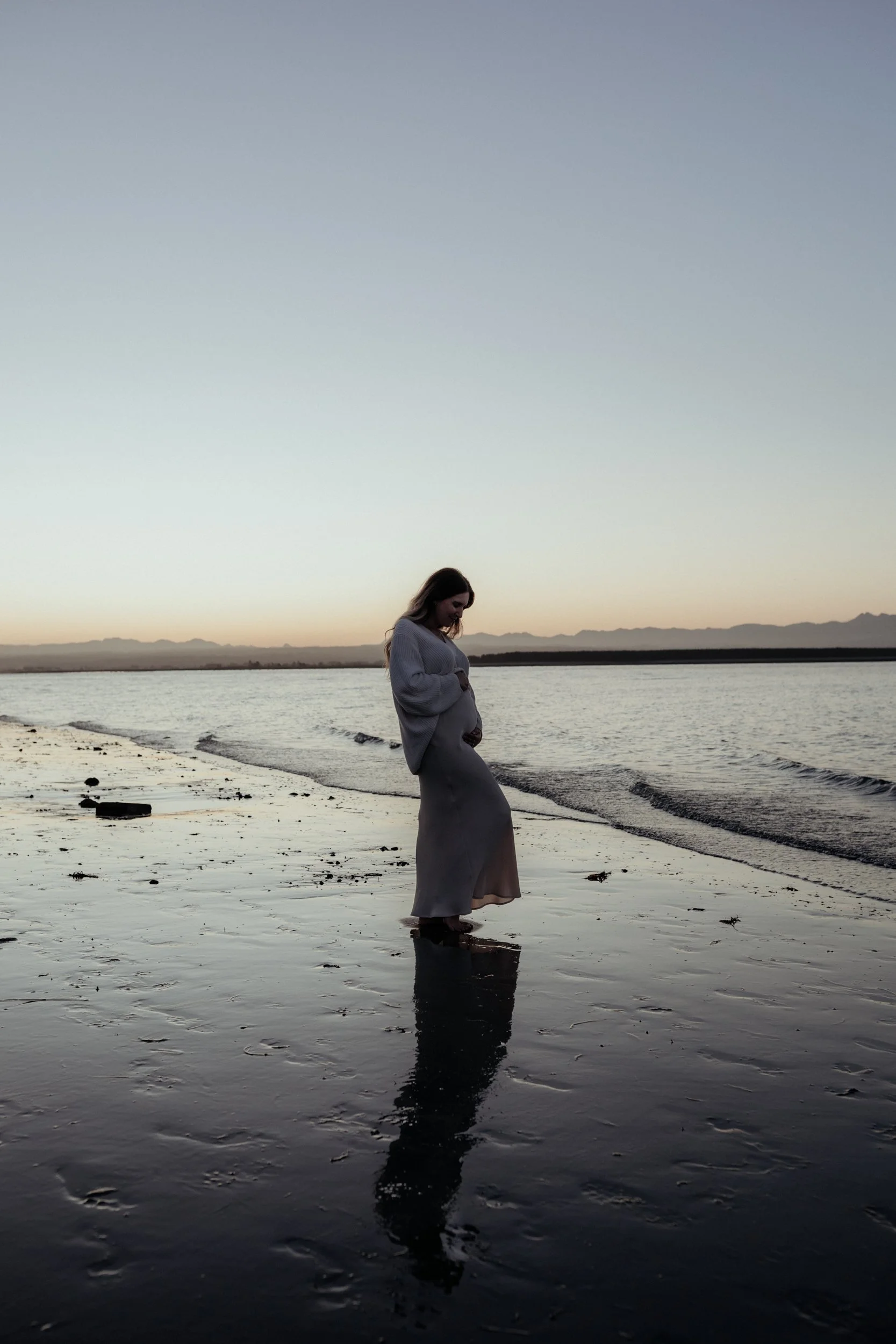 Expectant mother holds pregnant belly at sunset maternity photoshoot at Nelson beach.