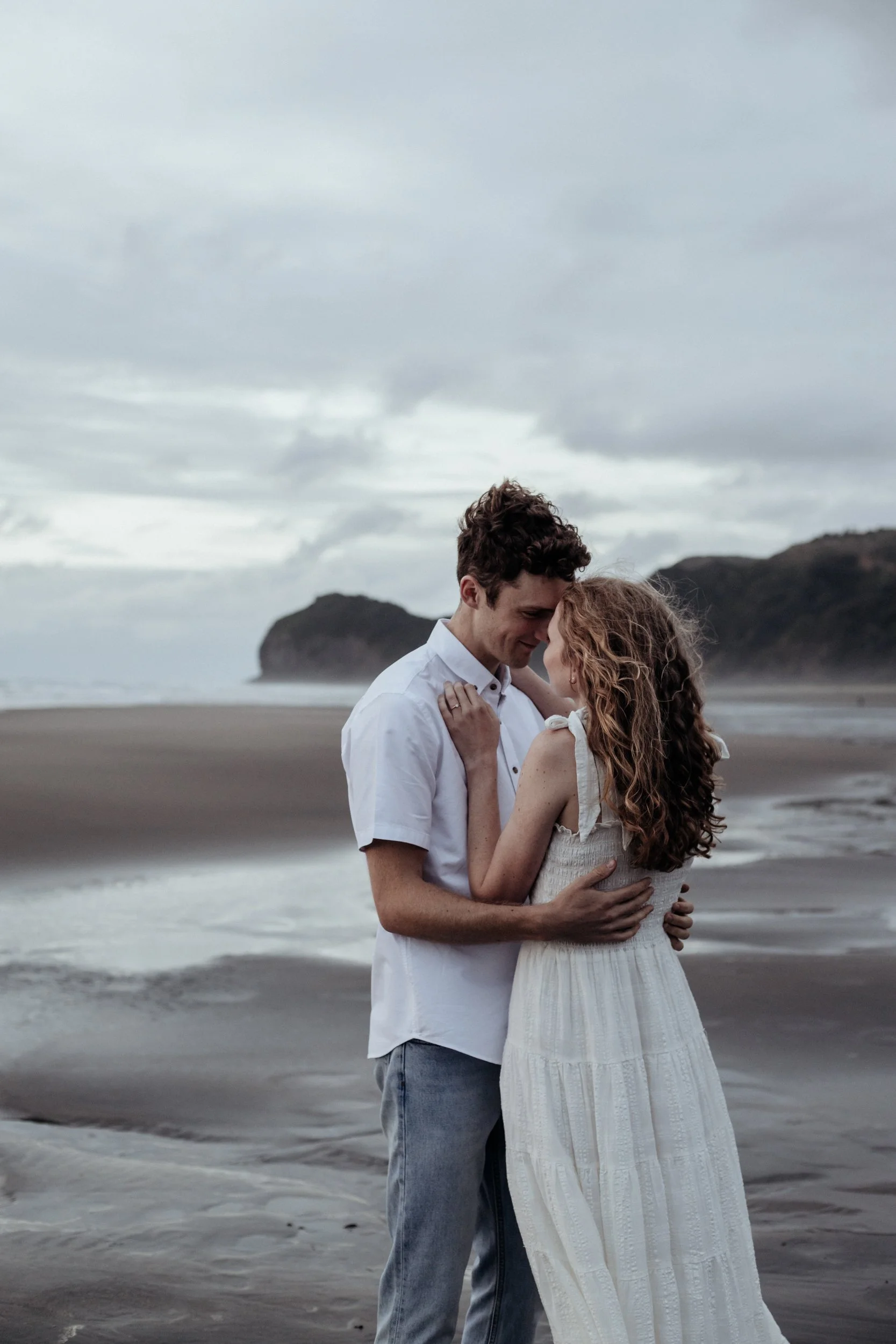 Couple embracing during sunset engagement session at Piha Beach.