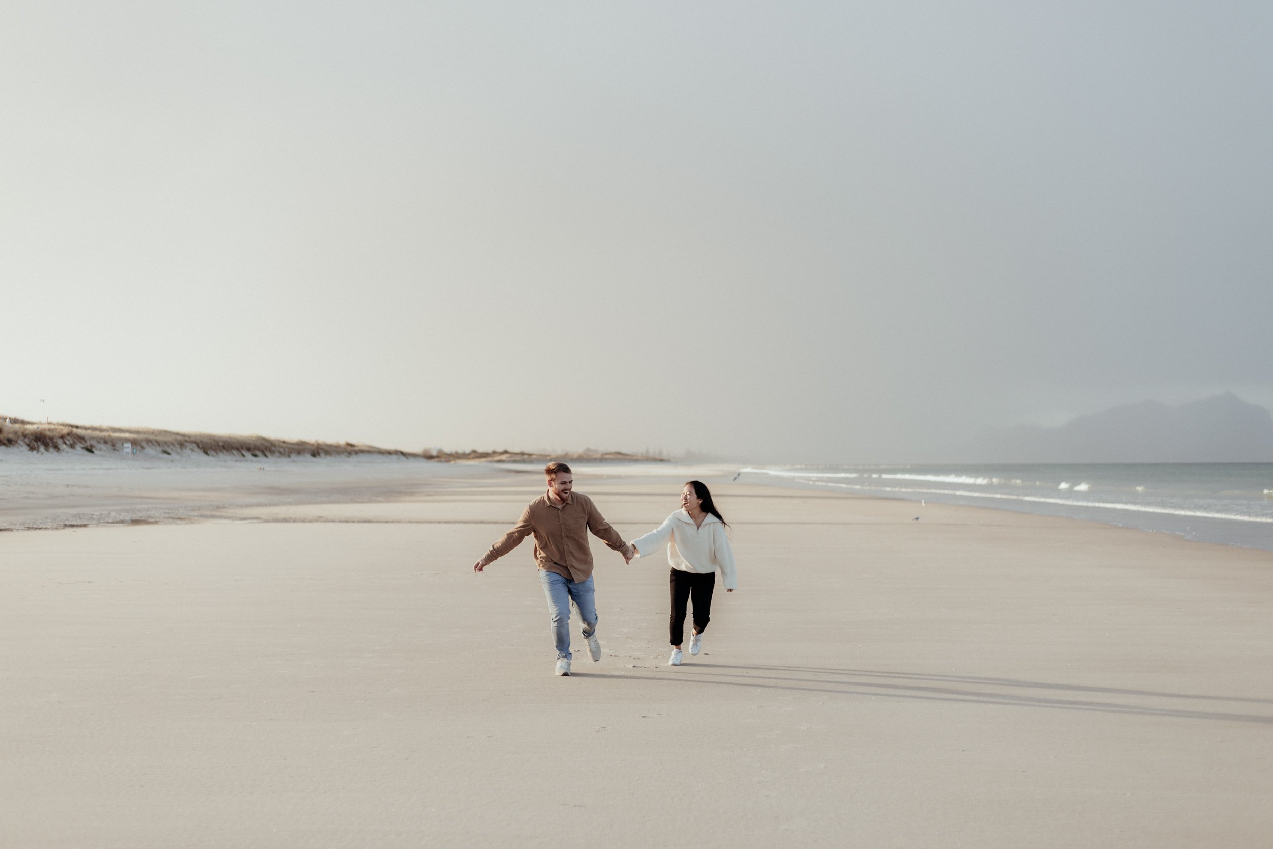 Newly engaged couple running on beach during a couple session in Whangarei at golden hour.