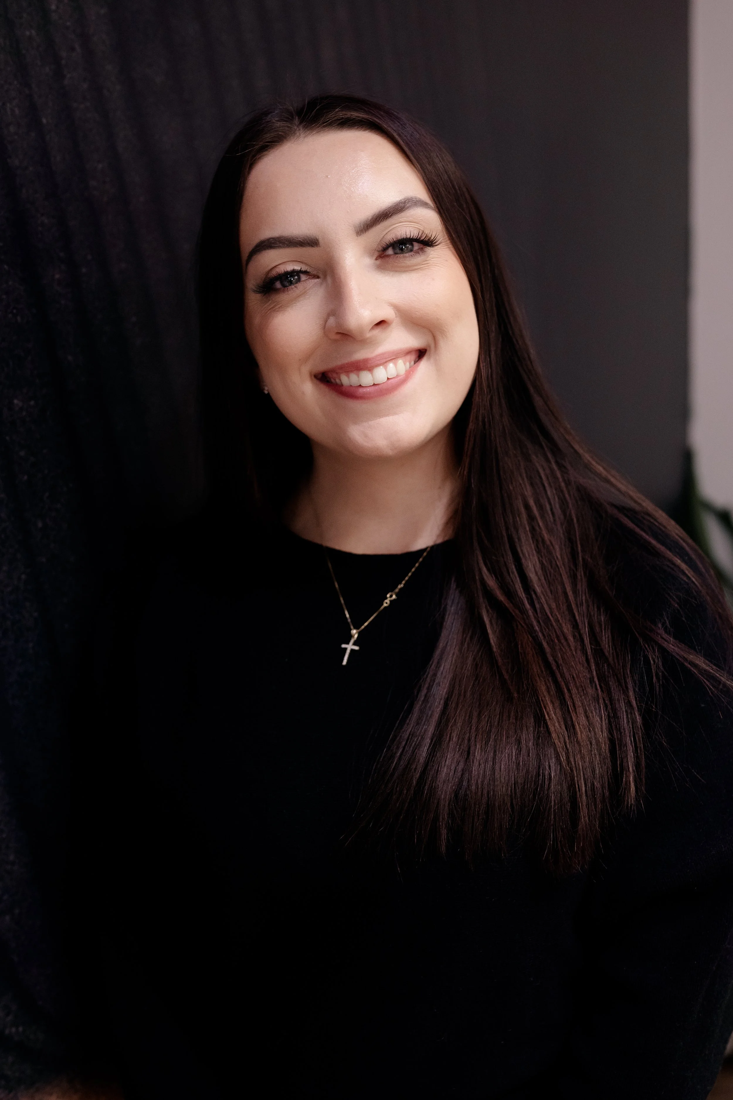 A young woman with long brown hair smiling and wearing a black top and a cross necklace.