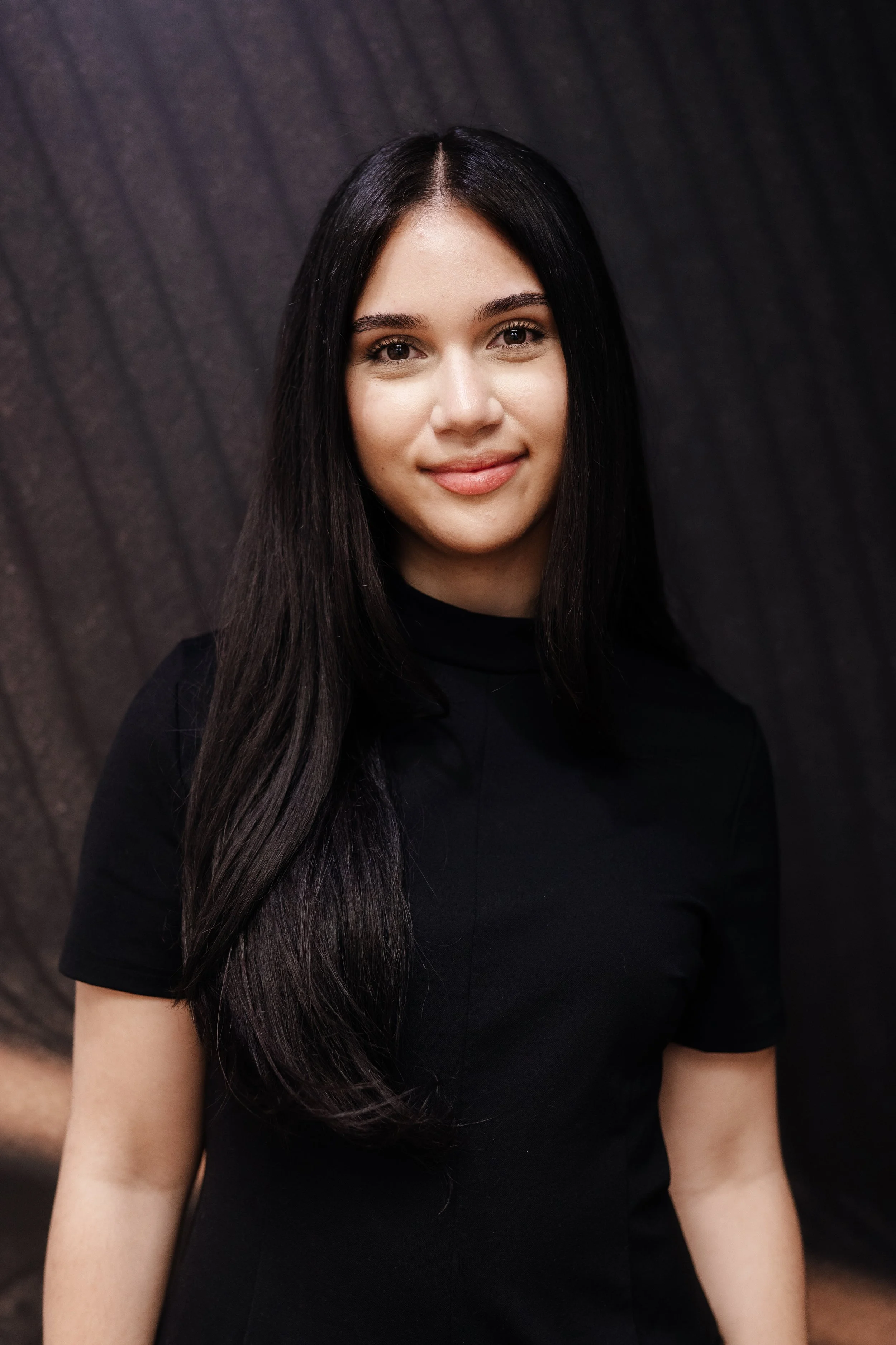 A young woman with long dark hair smiling at the camera, wearing a black top, with a dark textured background.