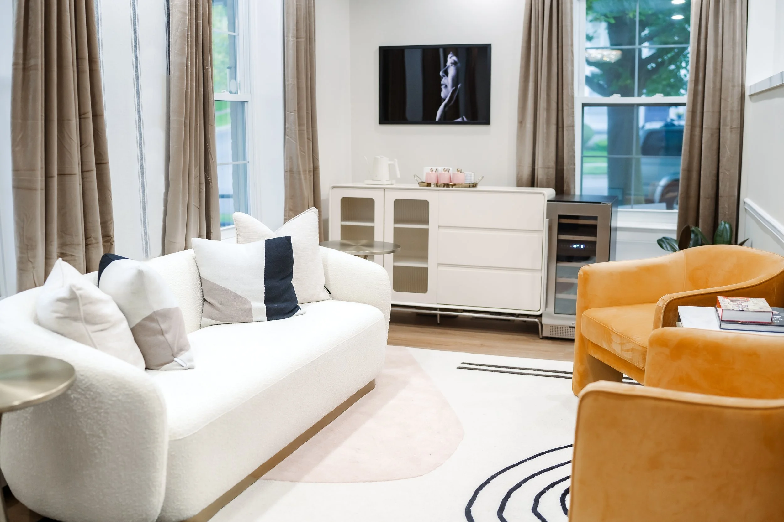 Living room with white sofa, black-and-white pillows, orange chairs, beige curtains, and a wall-mounted television.