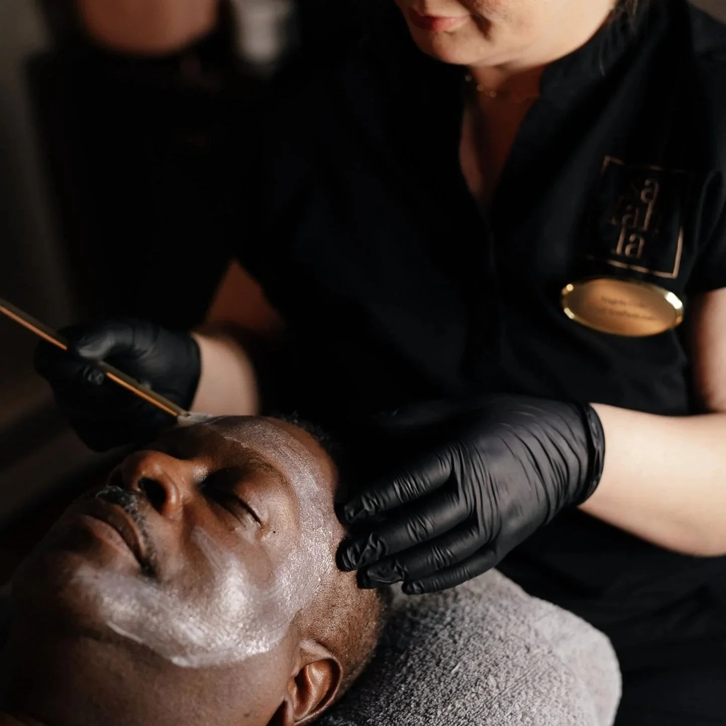 A person receiving a skincare facial treatment lying down with eyes closed, while a professional in black gloves applies a skincare product or treatment to their face.