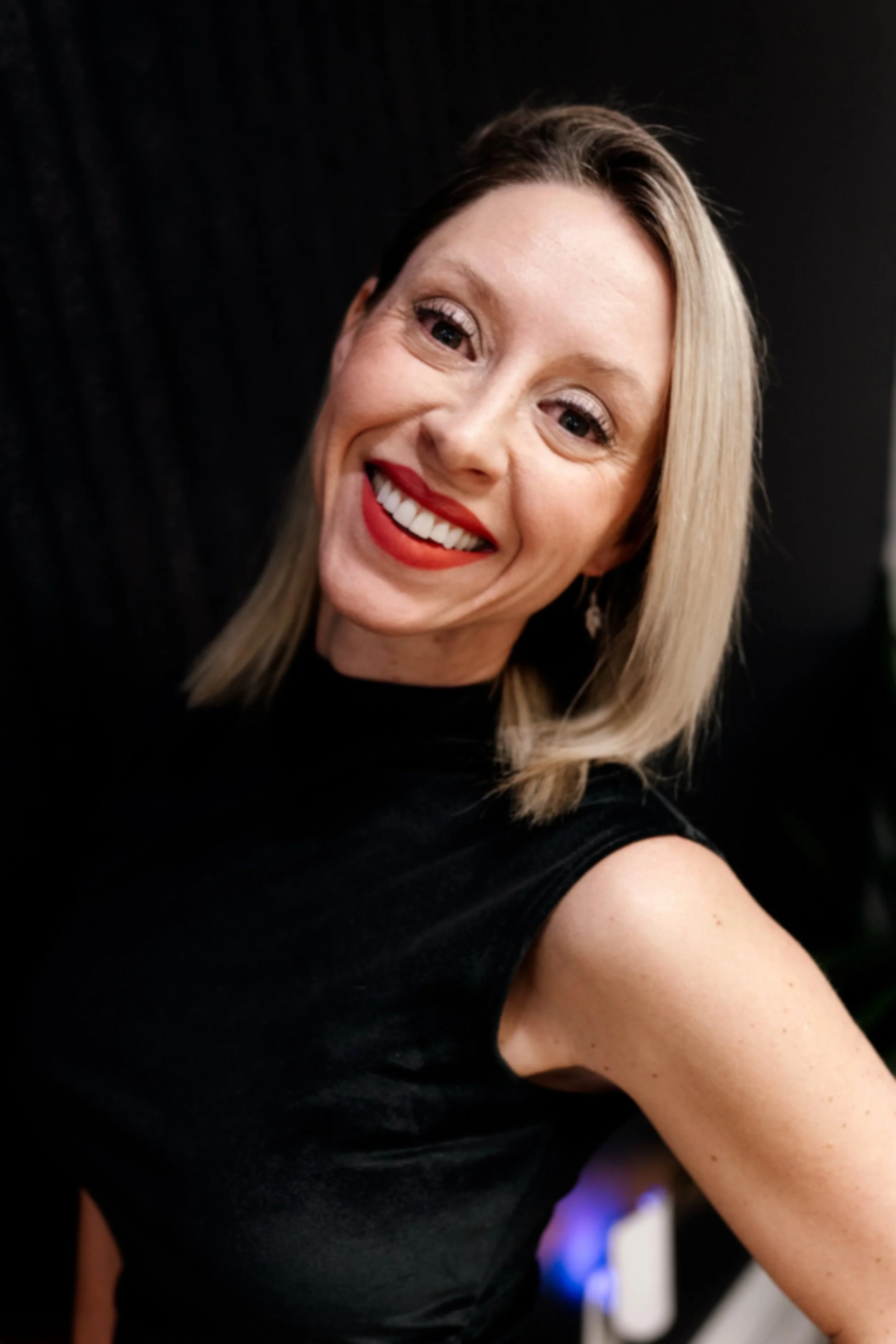 A woman smiling with red lipstick, blonde hair, wearing a black sleeveless top, and earrings, posing against a dark background.
