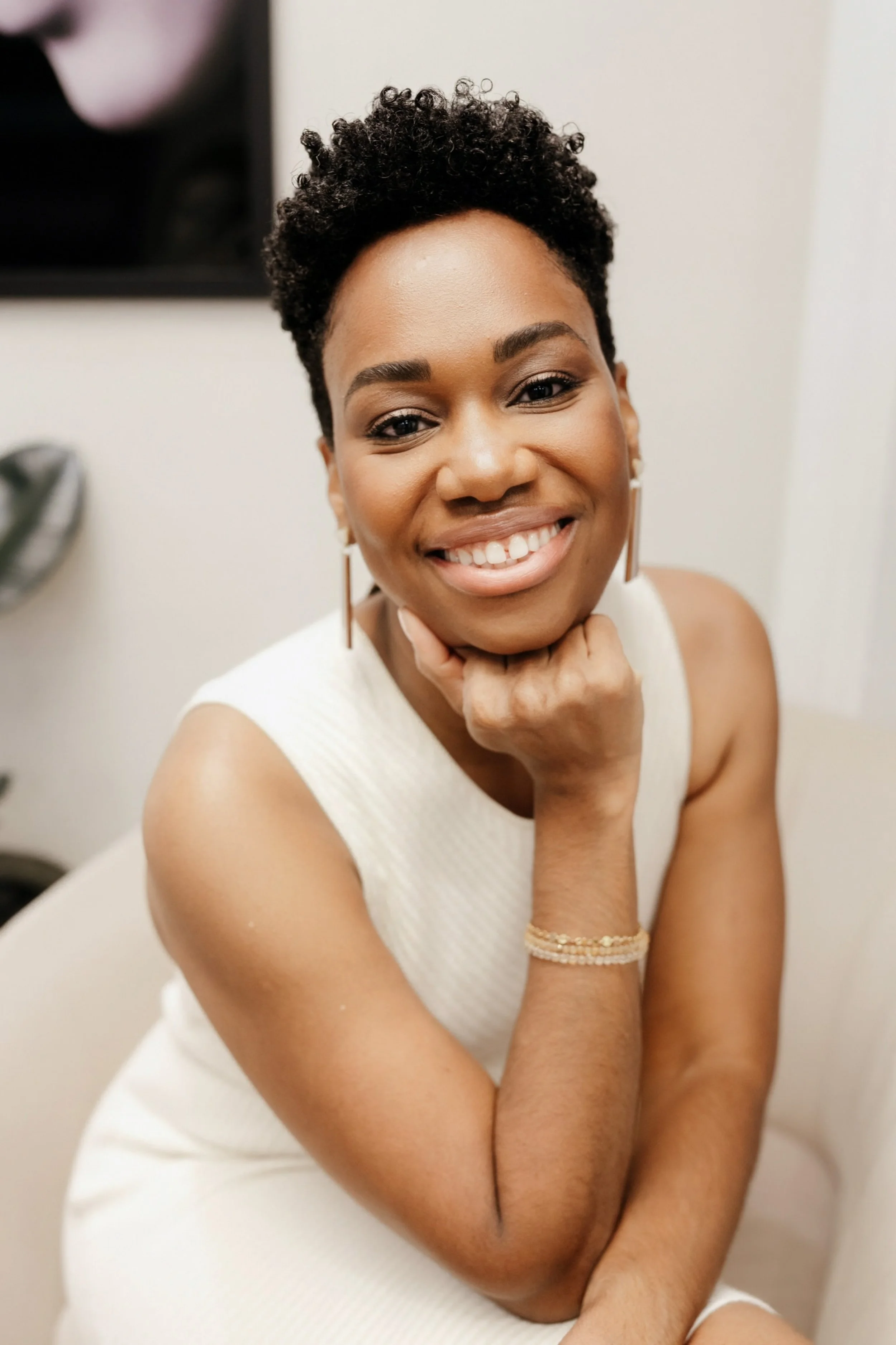 A smiling woman with short curly black hair, wearing a sleeveless white dress and gold jewelry, sitting with her chin resting on her hand in a well-lit room.