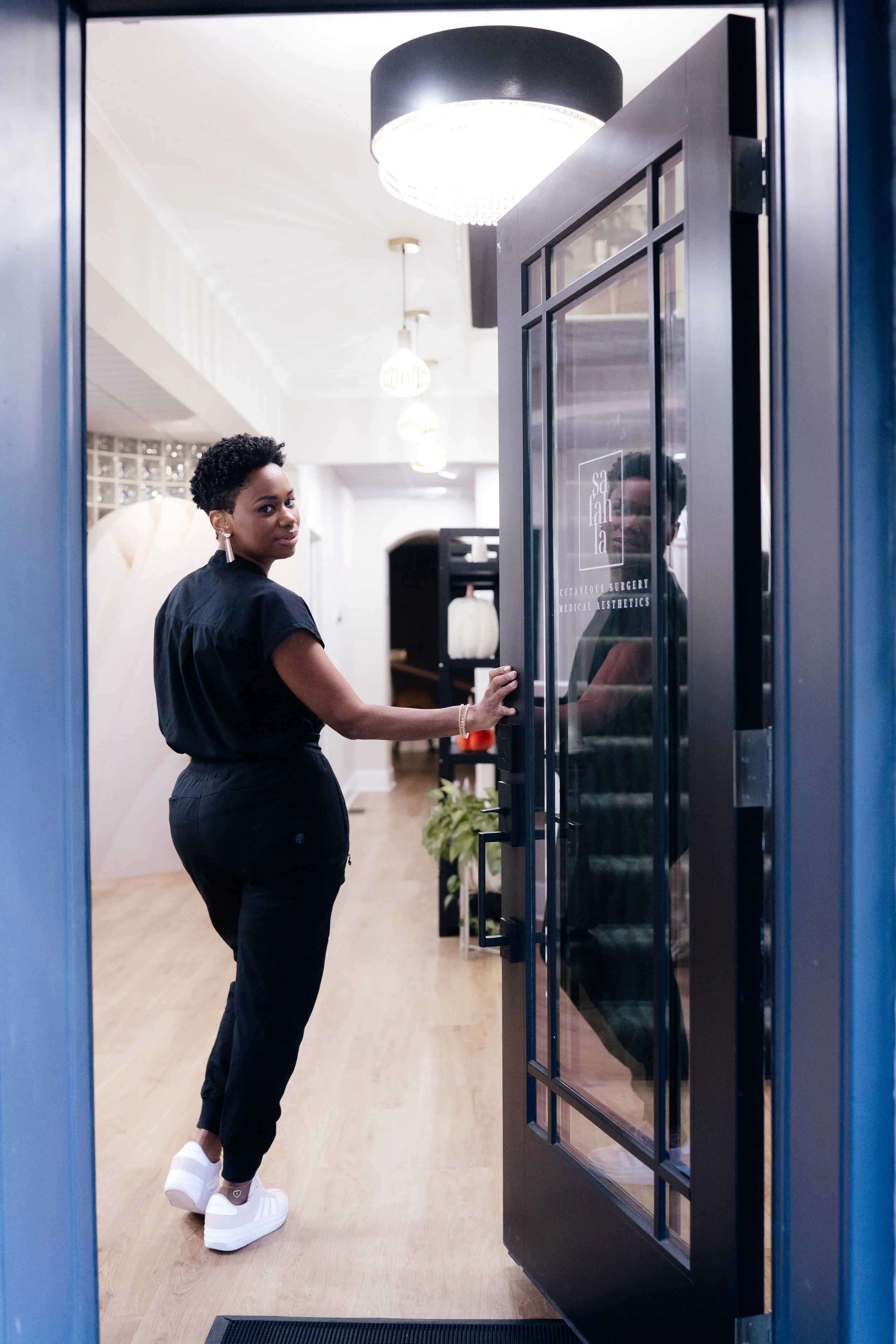 Woman opening glass door to a medical aesthetics office, interior with warm lighting, modern decor, and wooden flooring.