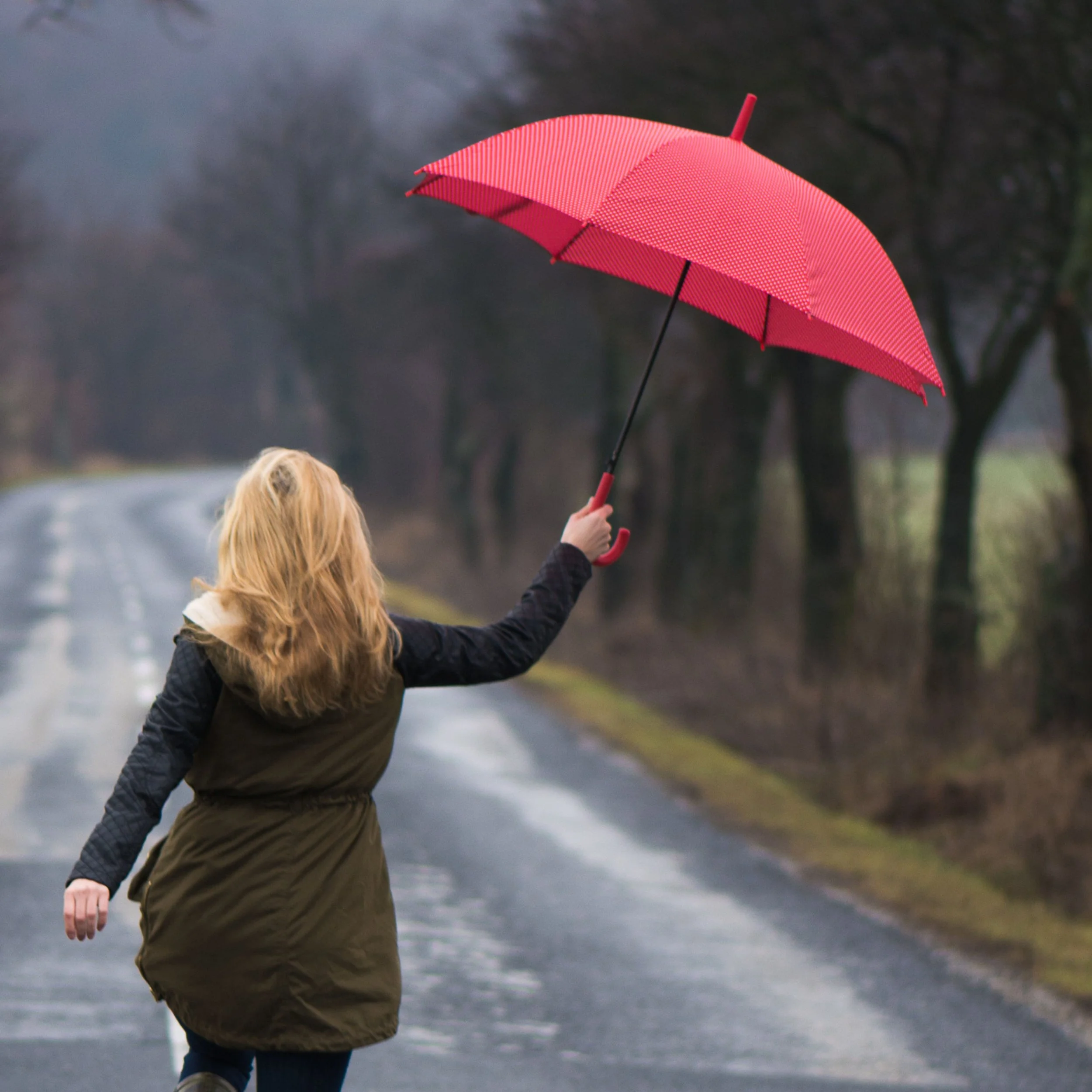 Woman with blond hair holding a pink umbrella as she walks down a wet, empty country road on a rainy day.
