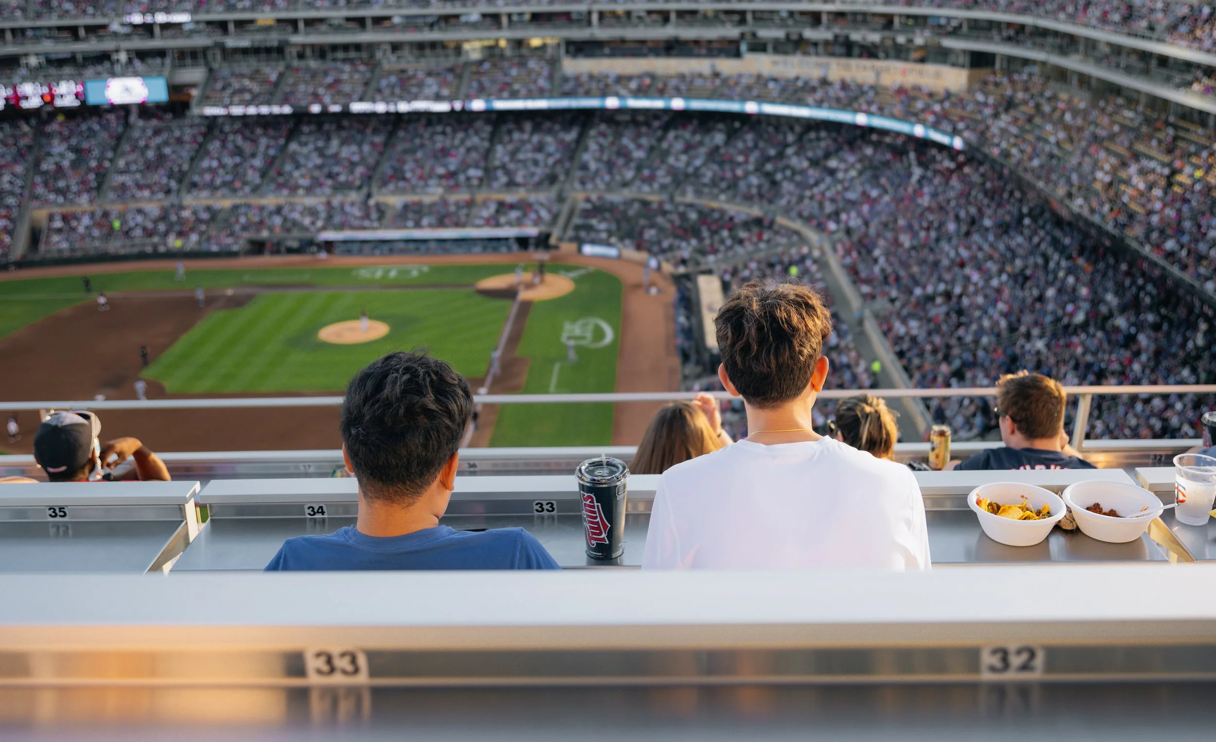 People watching a baseball game from stadium seats with bowls of food and drink on a ledge in the foreground.