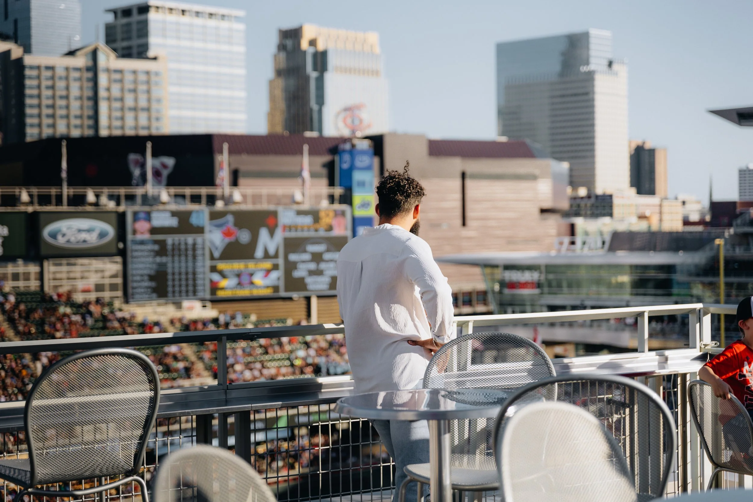 A man in a white shirt standing on a balcony at a baseball stadium, overlooking the field and scoreboard.