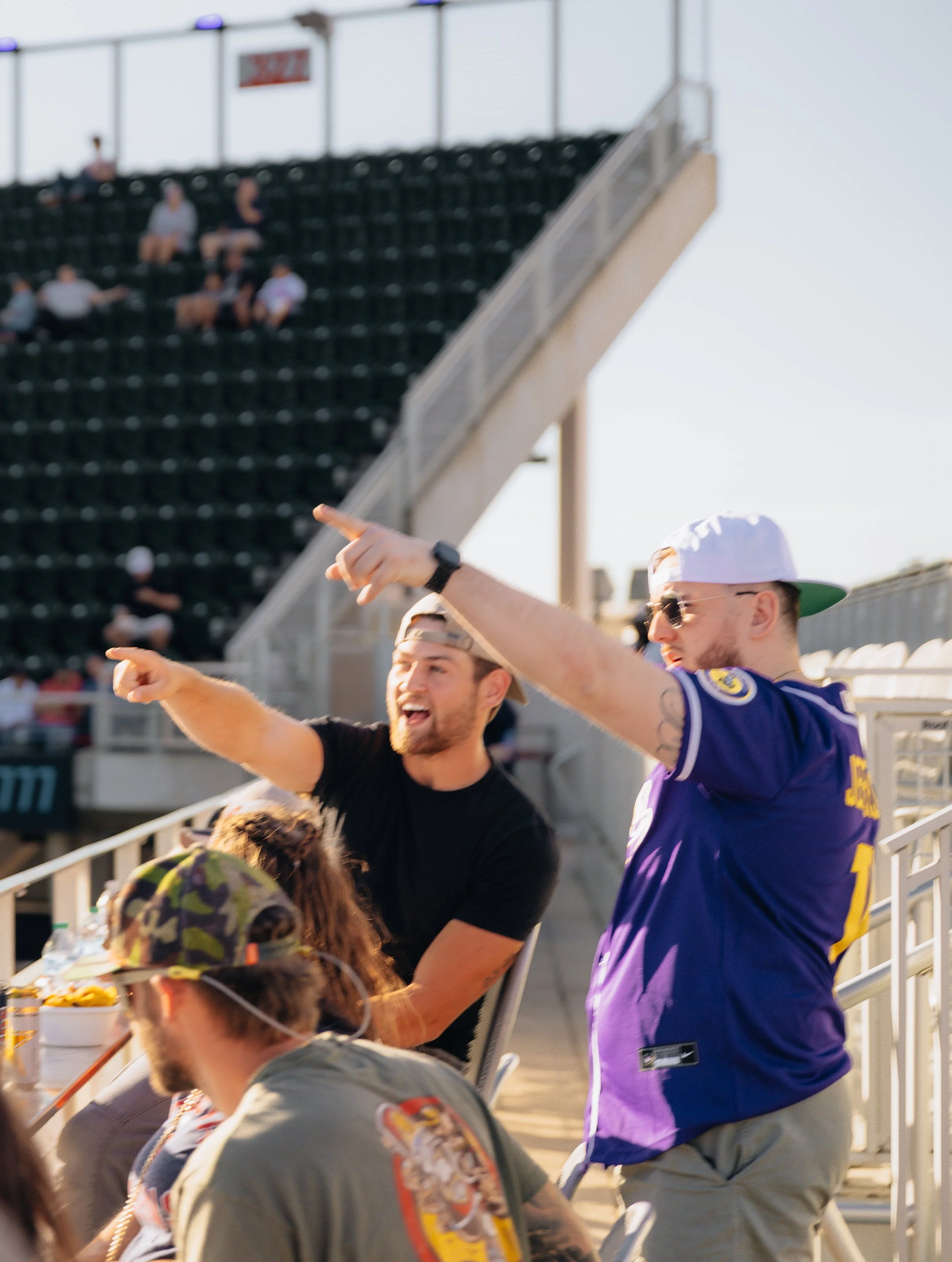 Two men at a stadium, one in a black T-shirt and the other in a purple sports jersey, are pointing and smiling, engaging with spectators seated nearby. Others are watching from the stands.