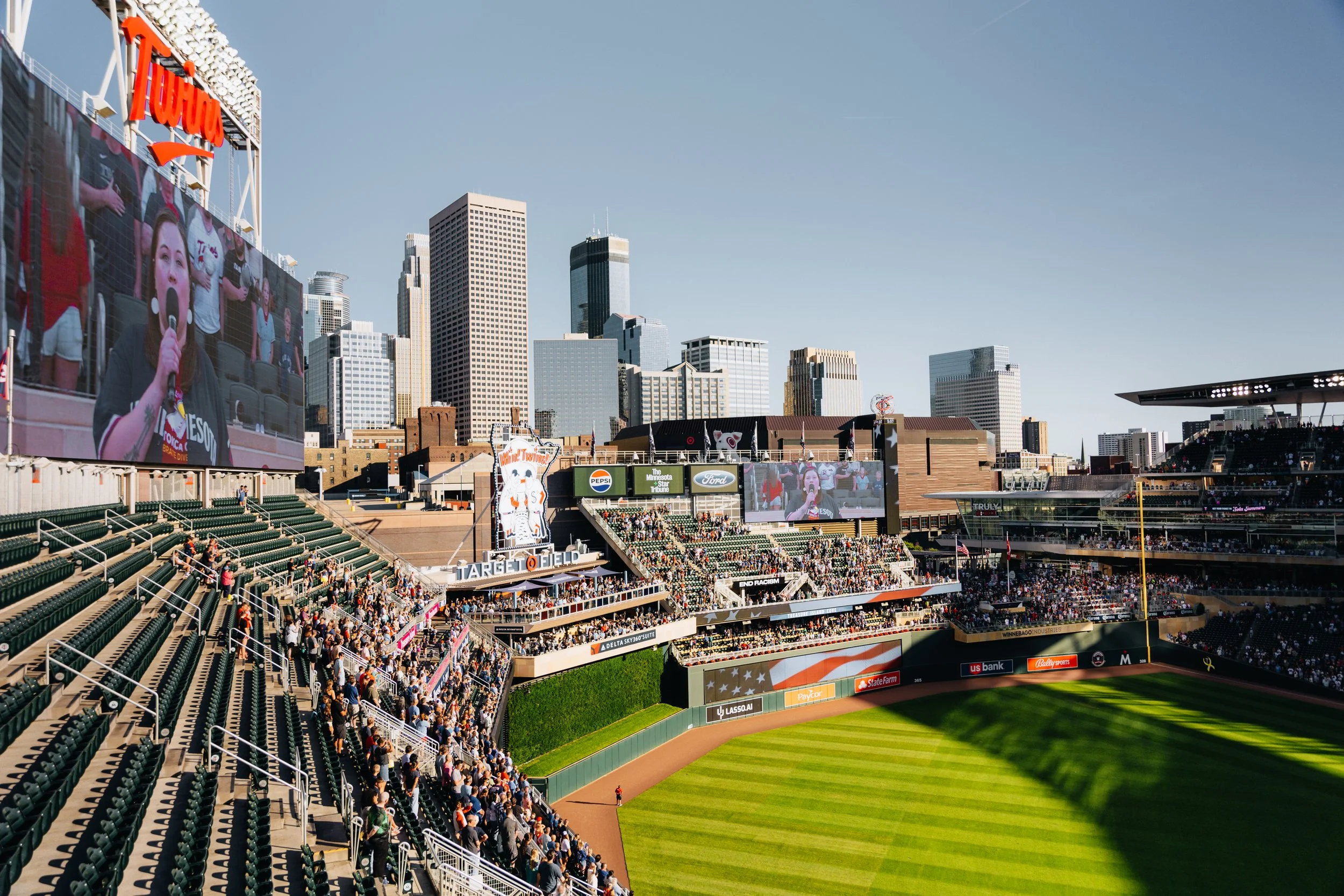 A baseball stadium filled with spectators and a large digital scoreboard showing a woman singing with a microphone, with a city skyline in the background.