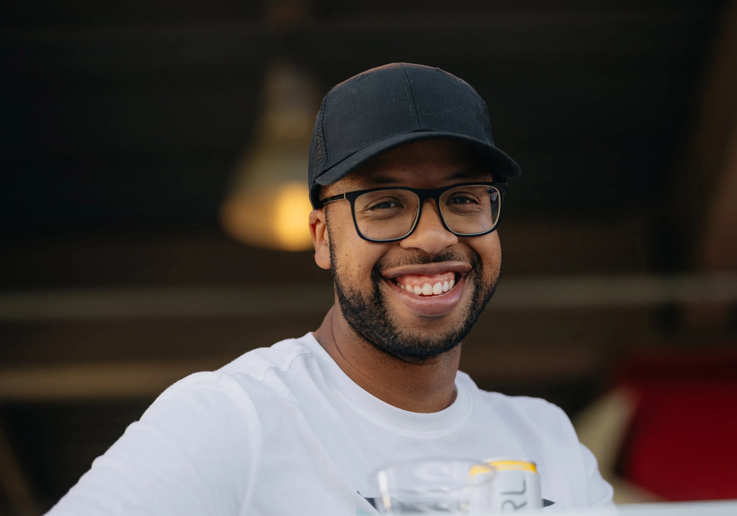 Smiling man wearing glasses, a black cap, and a white T-shirt, in an indoor setting with a blurred background.