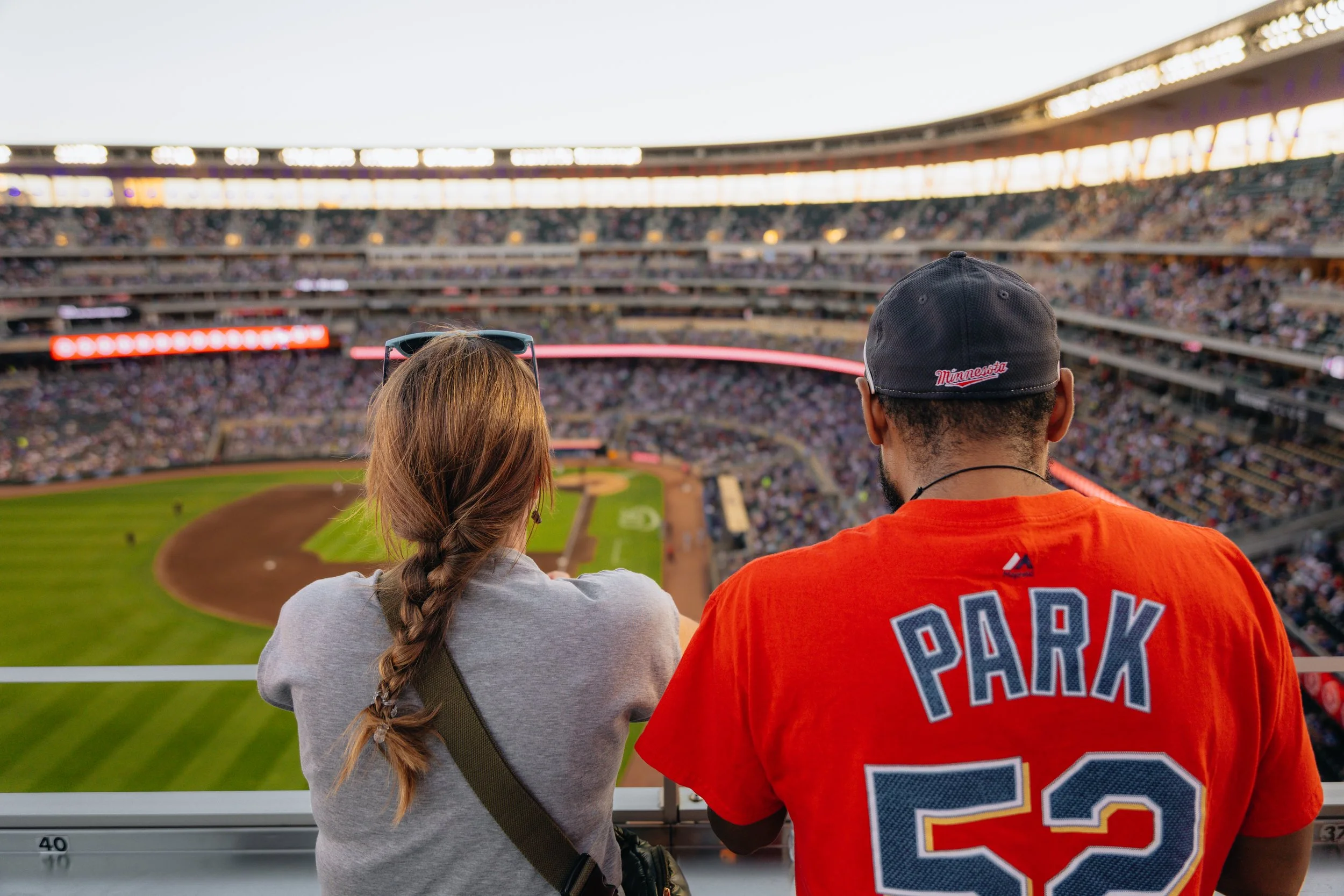 A woman and a man sitting in a stadium watching a baseball game, seen from behind, with the field and tiered seating filled with spectators in the background.