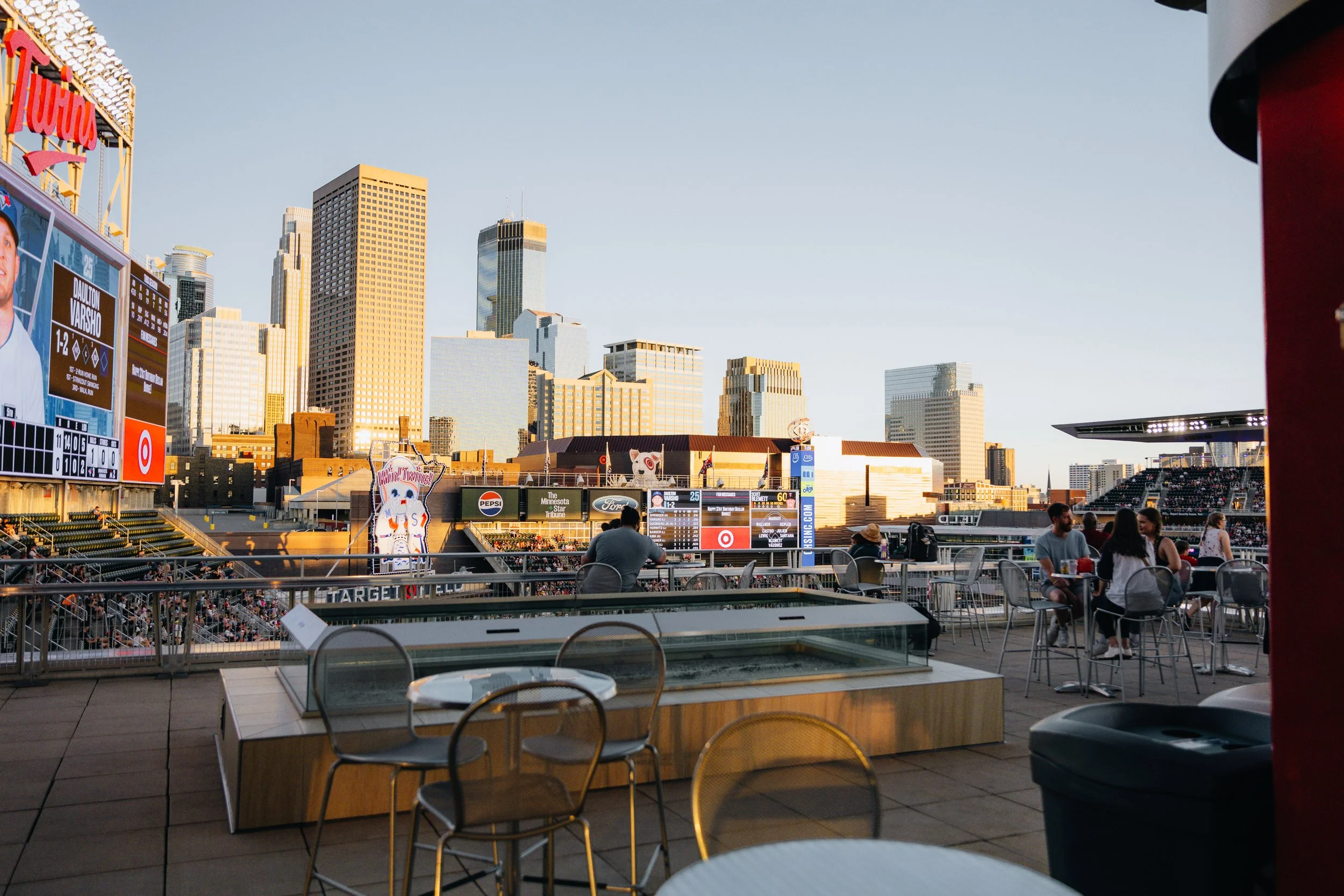 Tampa Bay Rays baseball stadium view from a outdoor seating area with a few people, city skyline in the background, and a large scoreboard displaying player information.