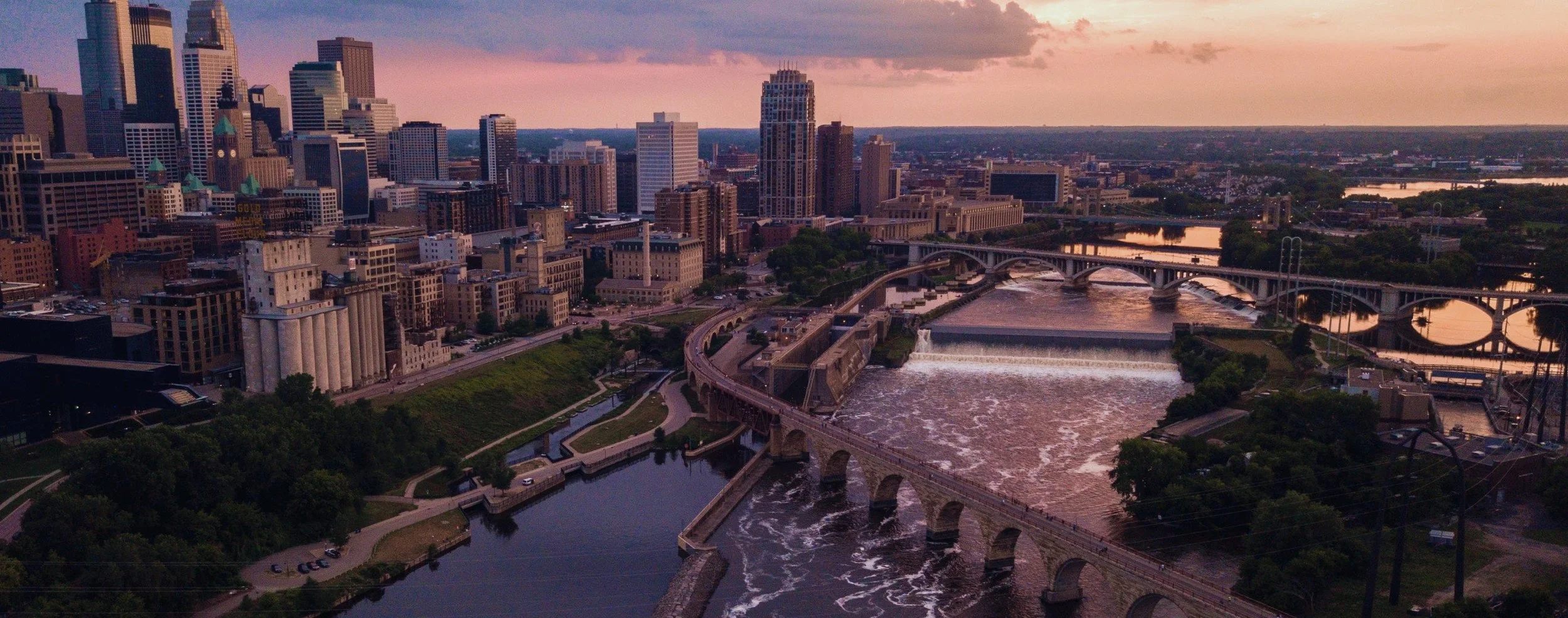 A panoramic view of the downtown minneapolis skyline at sunset with tall buildings, bridges, and a river with waterfalls.