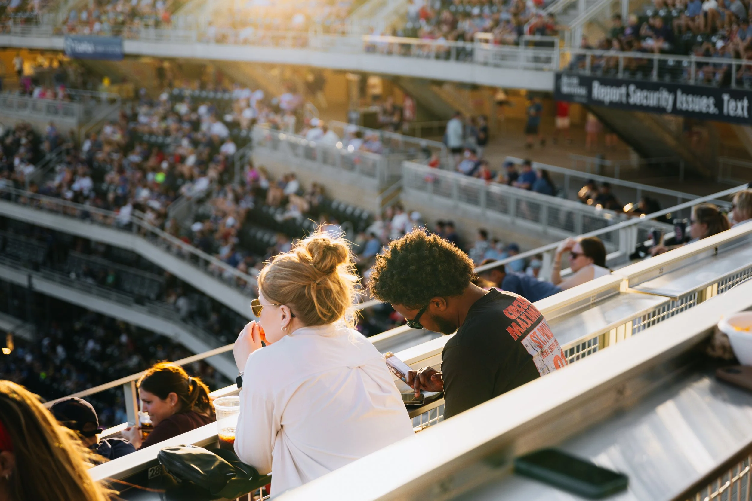 People sitting in stadium seats at an outdoor event, with many spectators in the background.