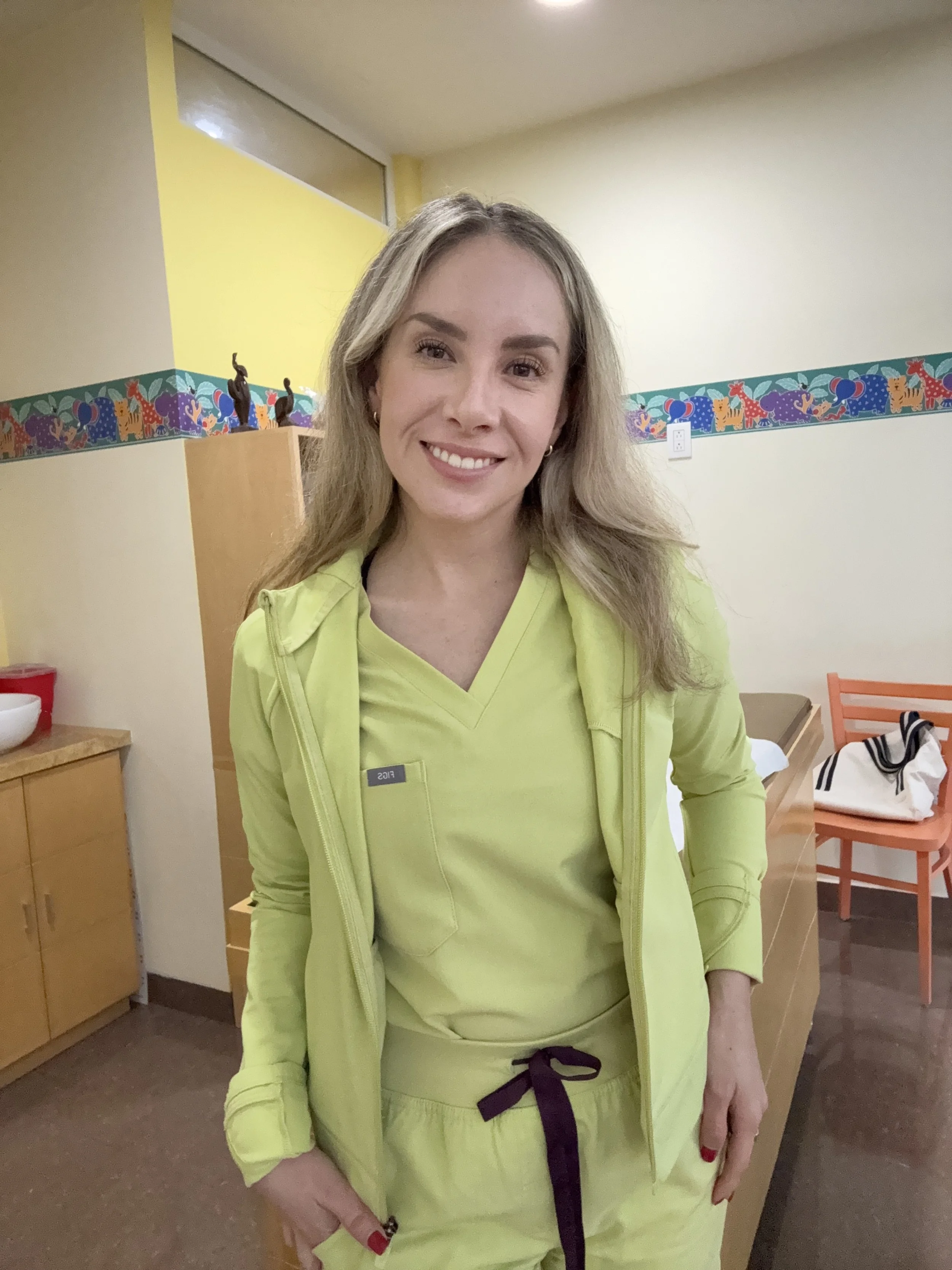 Mujer sonriendo, lleva uniforme de trabajo en un lugar con paredes color crema y decoraciones en movimiento de animales y globos, fondo con silla y mesa.