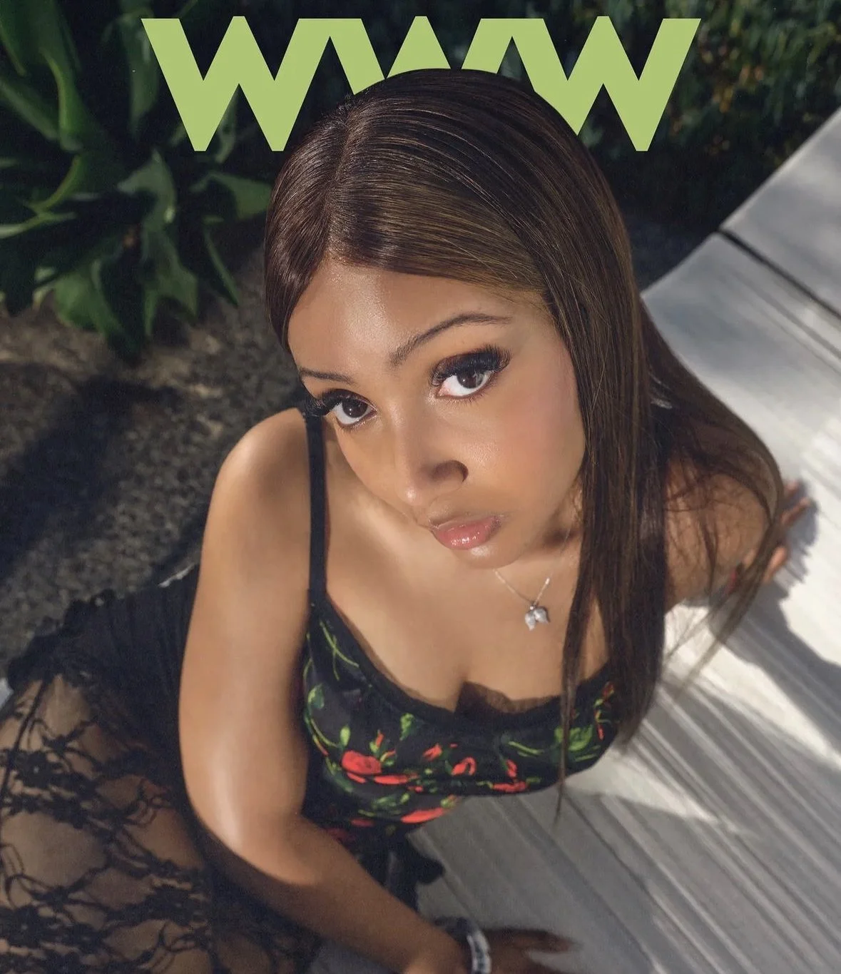 A woman with long brown hair and makeup looking up at the camera, wearing a black floral dress, standing on a wooden deck with greenery nearby, with the word "WOW" in large green letters at the top.