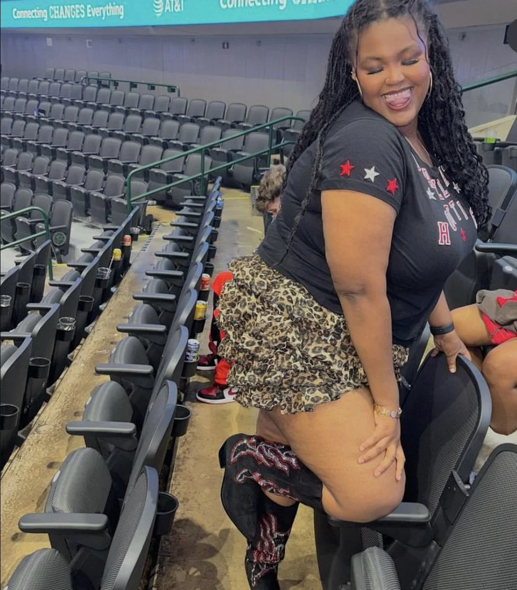 Smiling woman with long dreadlocks wearing a black shirt with red and white stars and a leopard print skirt, kneeling on one knee on a stadium seat, in an arena with empty black seats surrounding her.