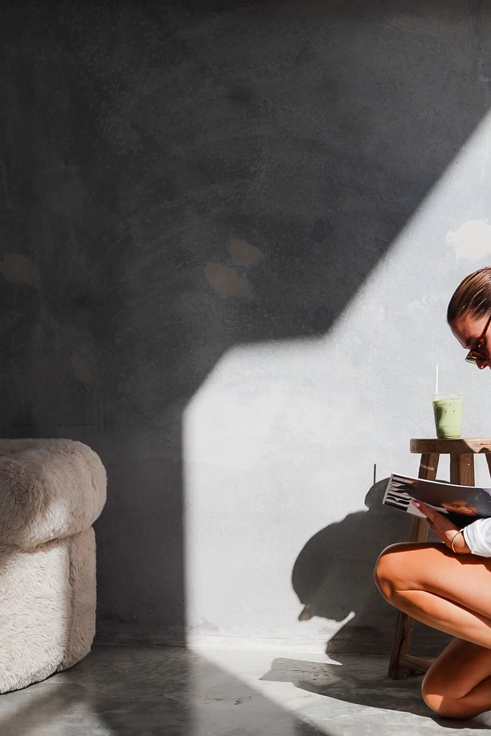 A woman kneeling on the floor reading a magazine next to a small wooden table with a green drink on it, with sunlight casting shadows on a gray wall.