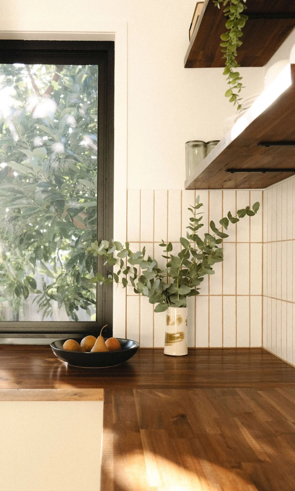 A kitchen countertop with a black bowl of pears, a vase with green eucalyptus branches, and a window with greenery outside.