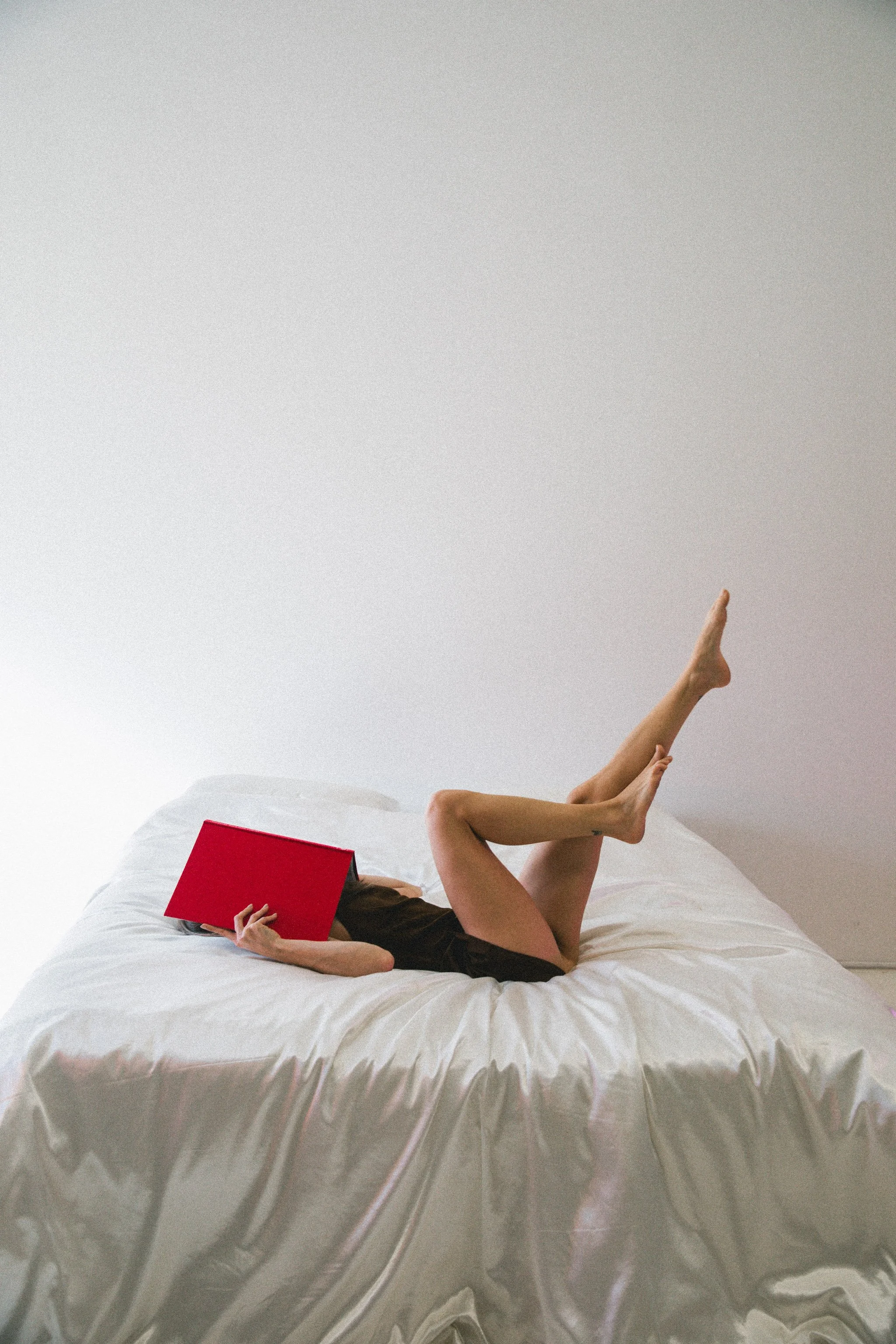 A woman lying on a bed with a metallic blanket, reading a red book with her face obscured by the book, and her legs crossed in the air.