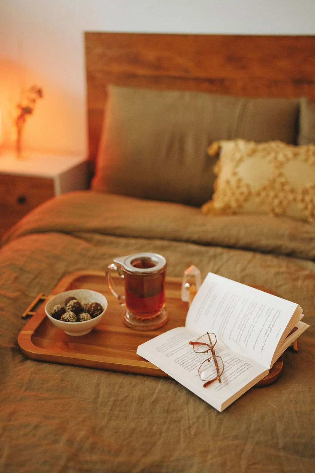 A bedroom with a wooden bed and beige pillows. A wooden tray on the bed holds a cup of tea, a bowl of chocolate truffles, a pair of reading glasses, an open book, and some small items. A bedside table with a lit lamp and a flower vase is visible in t