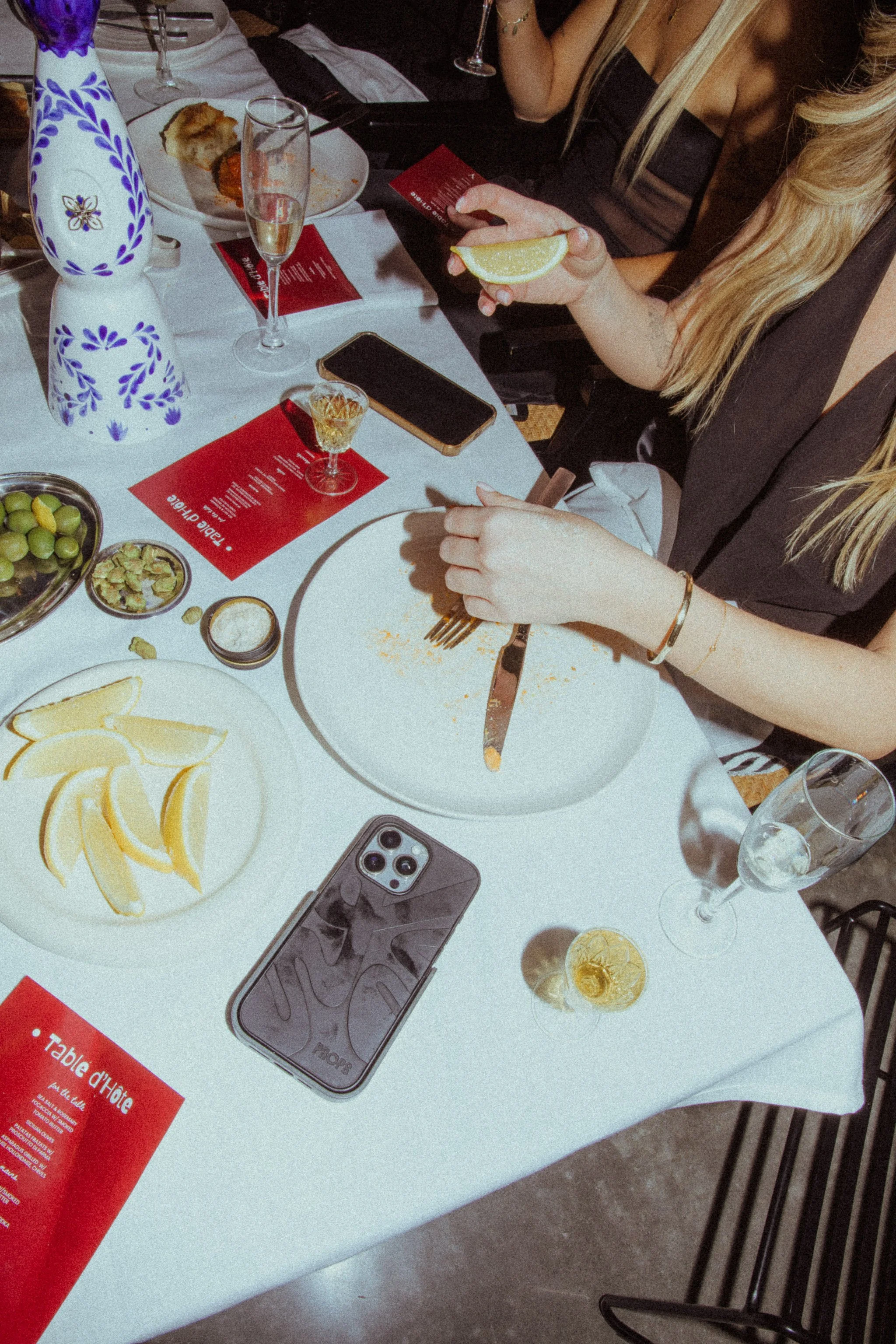 A woman sitting at a table with lemon wedges, a smartphone, drinking glasses, a plate with lemon slices, a bowl of grapes, and some small dishes. There are other people seated around, and the table has a white tablecloth.