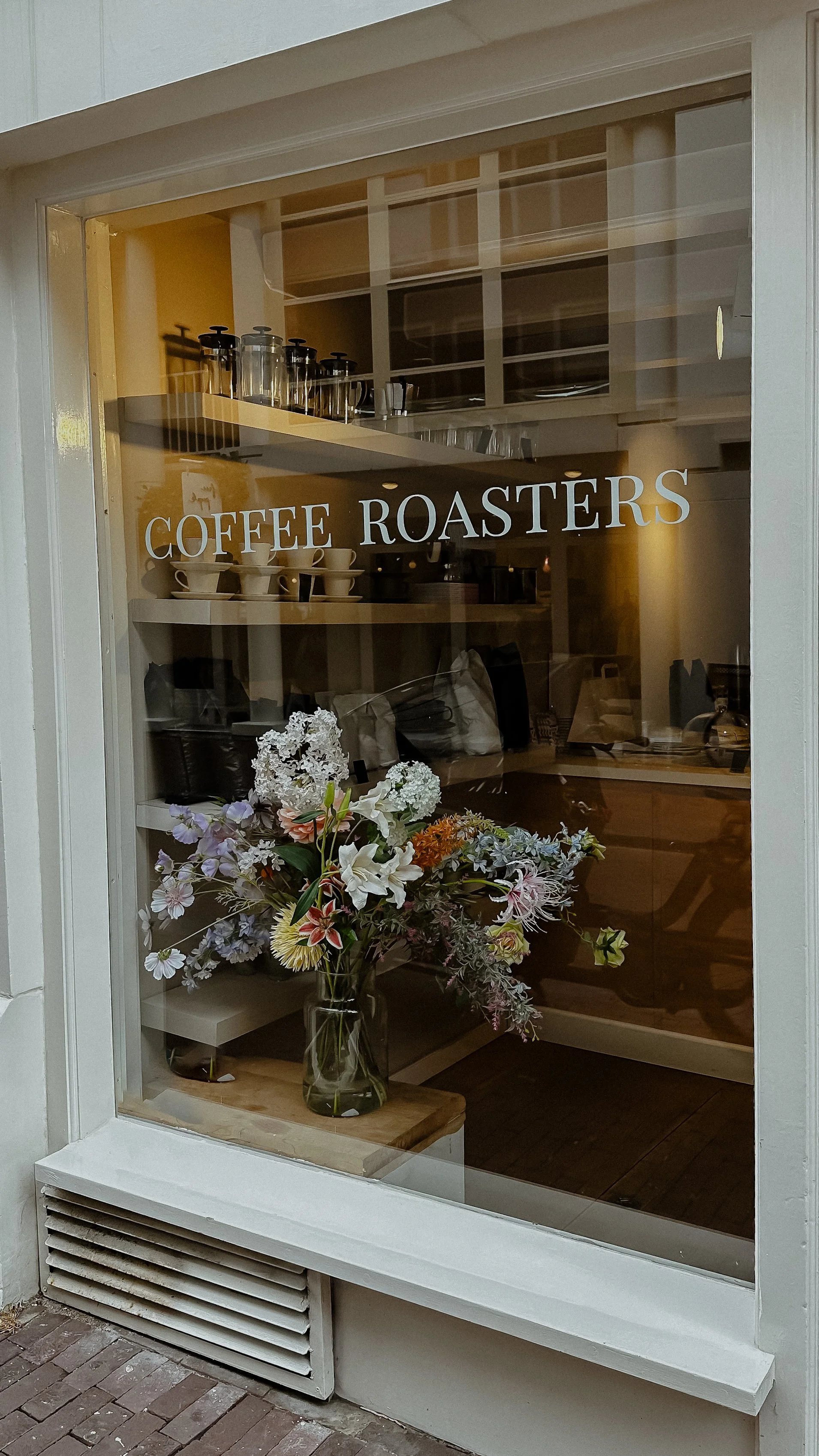 View through a cafe window showing a sign that reads 'Coffee Roasters,' a vase with a large bouquet of colorful flowers, and shelves with coffee equipment inside.