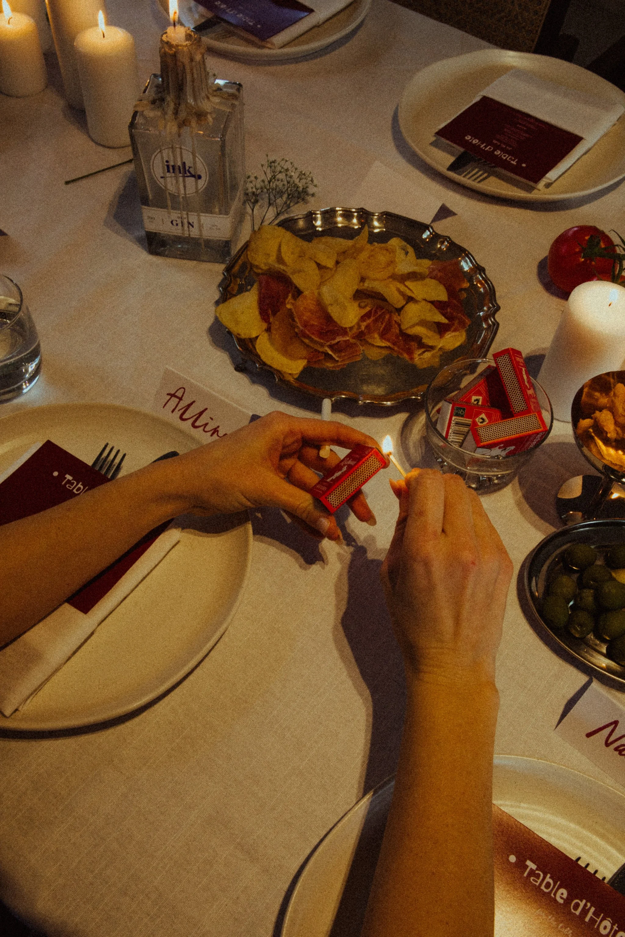 Person lighting a matchstick near a box of matches on a dinner table with snacks, candles, and a glass of gin.