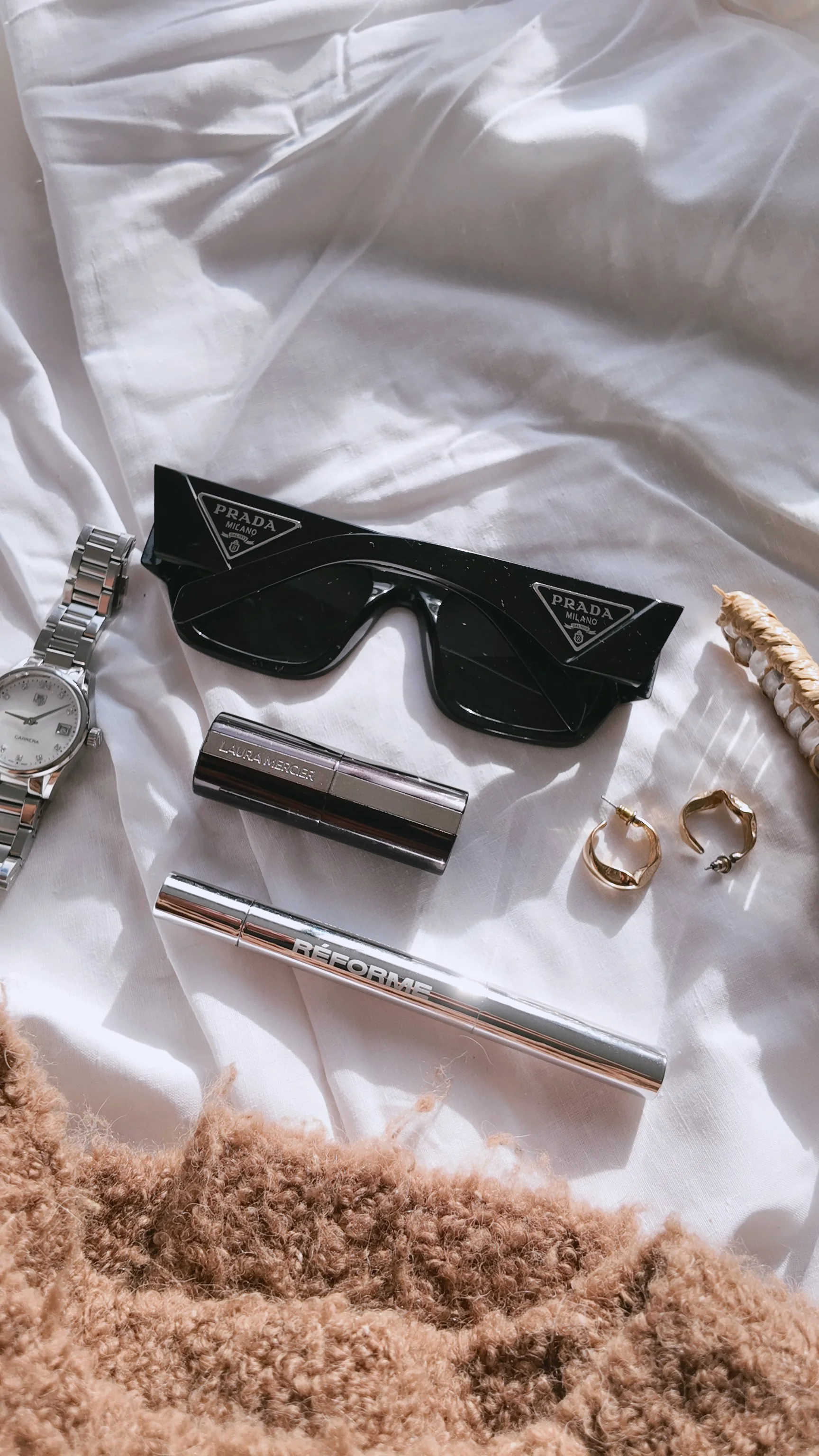 A collection of fashion accessories and cosmetics on a white fabric surface, including Prada sunglasses, a silver watch, Laura Mercier lipstick, Réponse perfume, and two gold rings, with a beige textured fabric in the foreground.