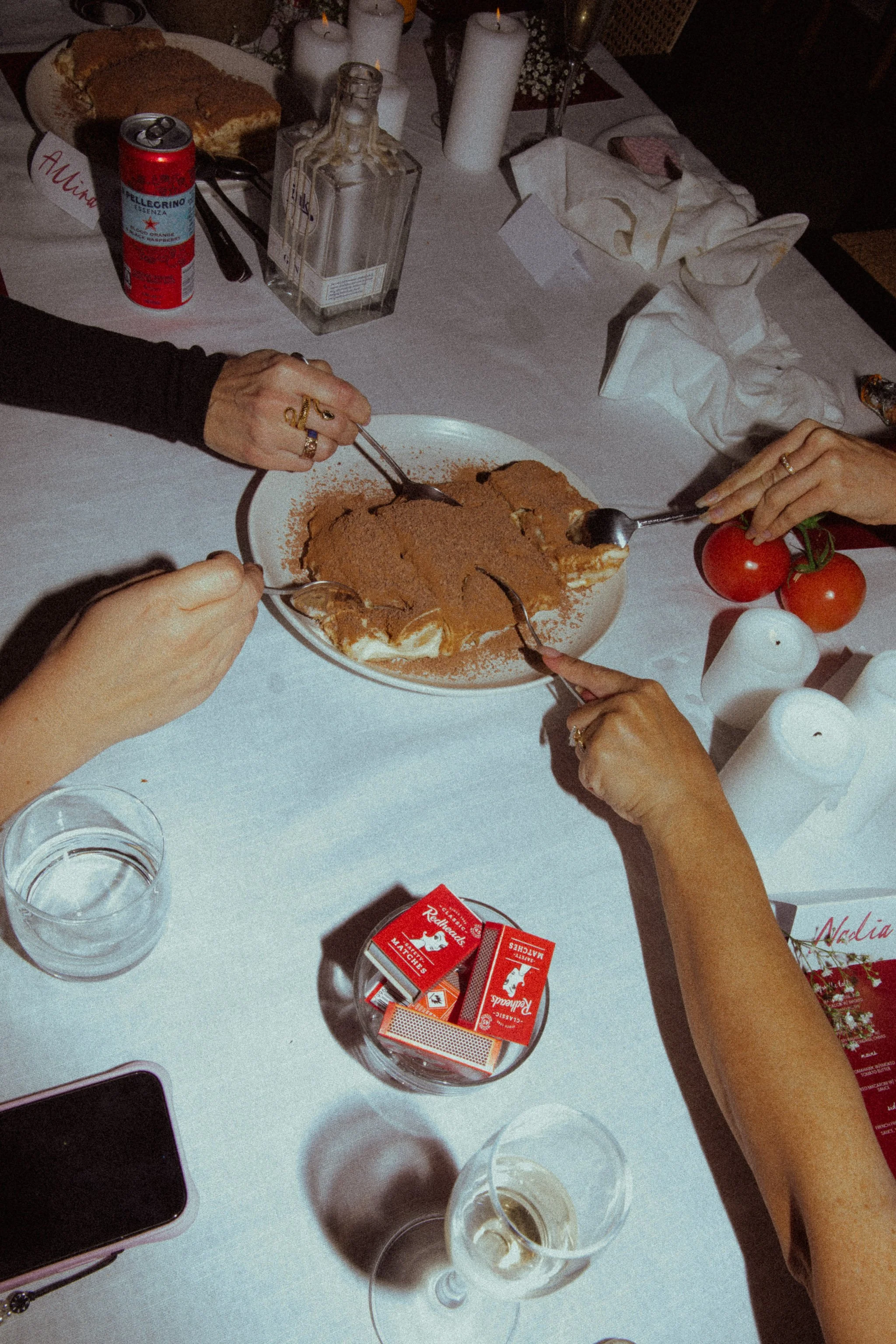 People sharing a tiramisu dessert at a dinner party with candles, wine glasses, and other tableware.