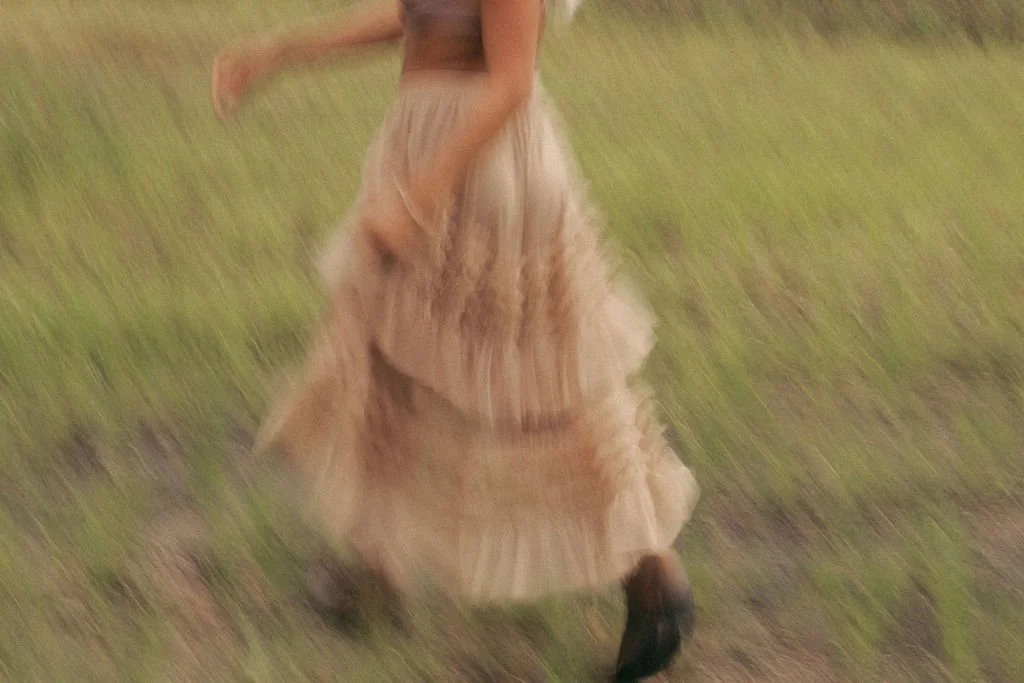 A person is walking outdoors on a grassy area wearing a long beige skirt and black shoes, captured in motion with a blurred effect.