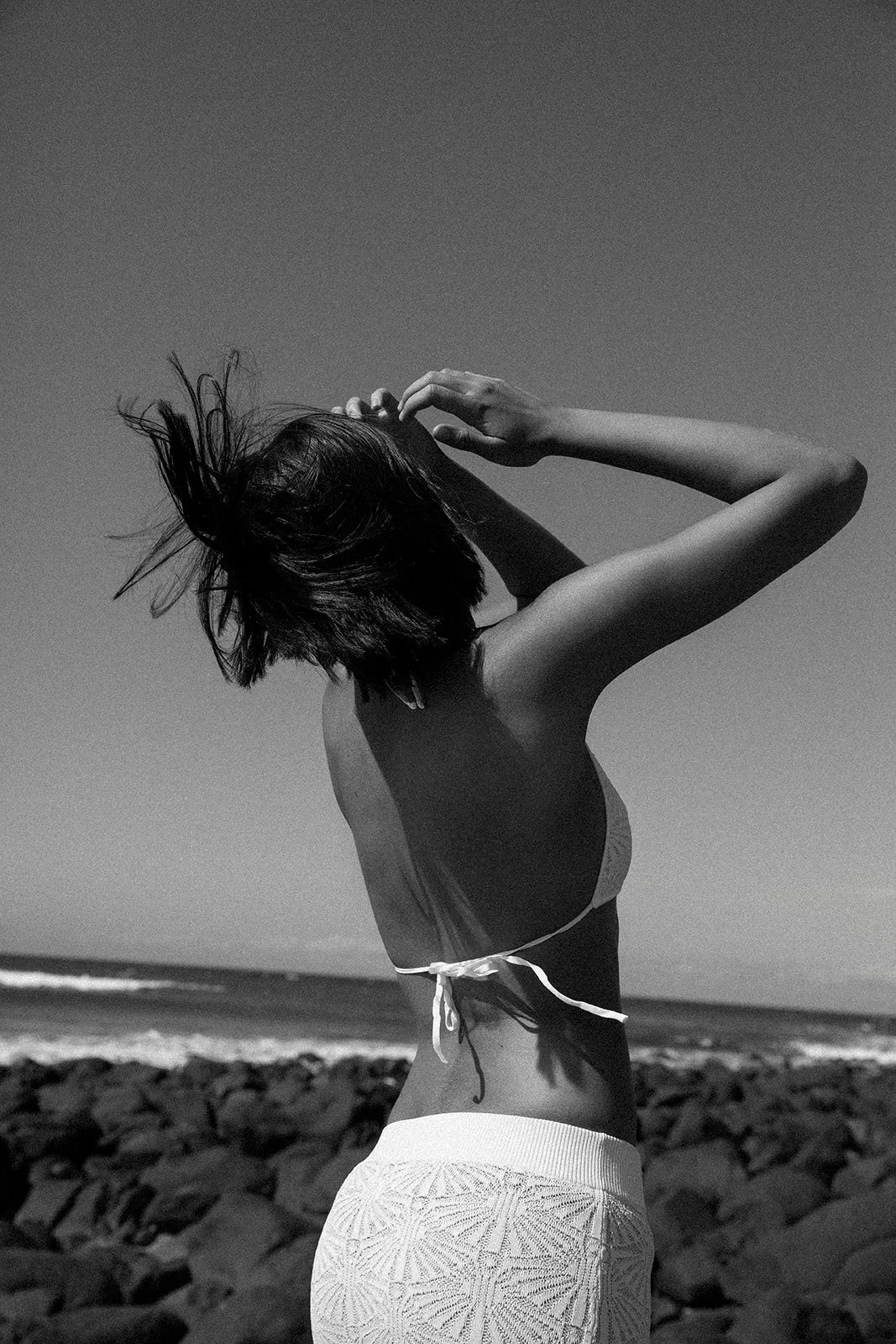 A woman in a bikini top and patterned skirt standing on a rocky beach with her hair blowing in the wind, facing away from the camera, in black and white.